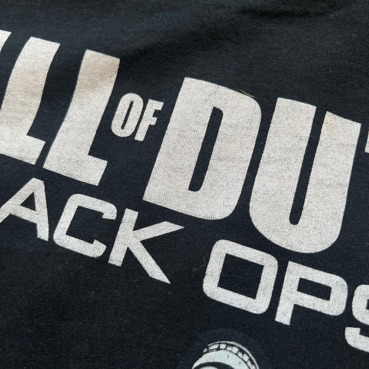 Call of Duty: Black Ops 2018 Black Tee is a black promotional T-shirt with a 2018 Activision Publishing licensed neck tag. The front features a large white and grey character graphic in armor holding a weapon, with the “Call of Duty Black Ops” logo printed above. The print spans most of the front from top to bottom.
