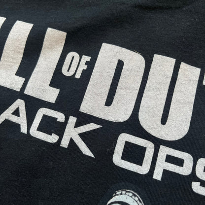 Call of Duty: Black Ops 2018 Black Tee is a black promotional T-shirt with a 2018 Activision Publishing licensed neck tag. The front features a large white and grey character graphic in armor holding a weapon, with the “Call of Duty Black Ops” logo printed above. The print spans most of the front from top to bottom.
