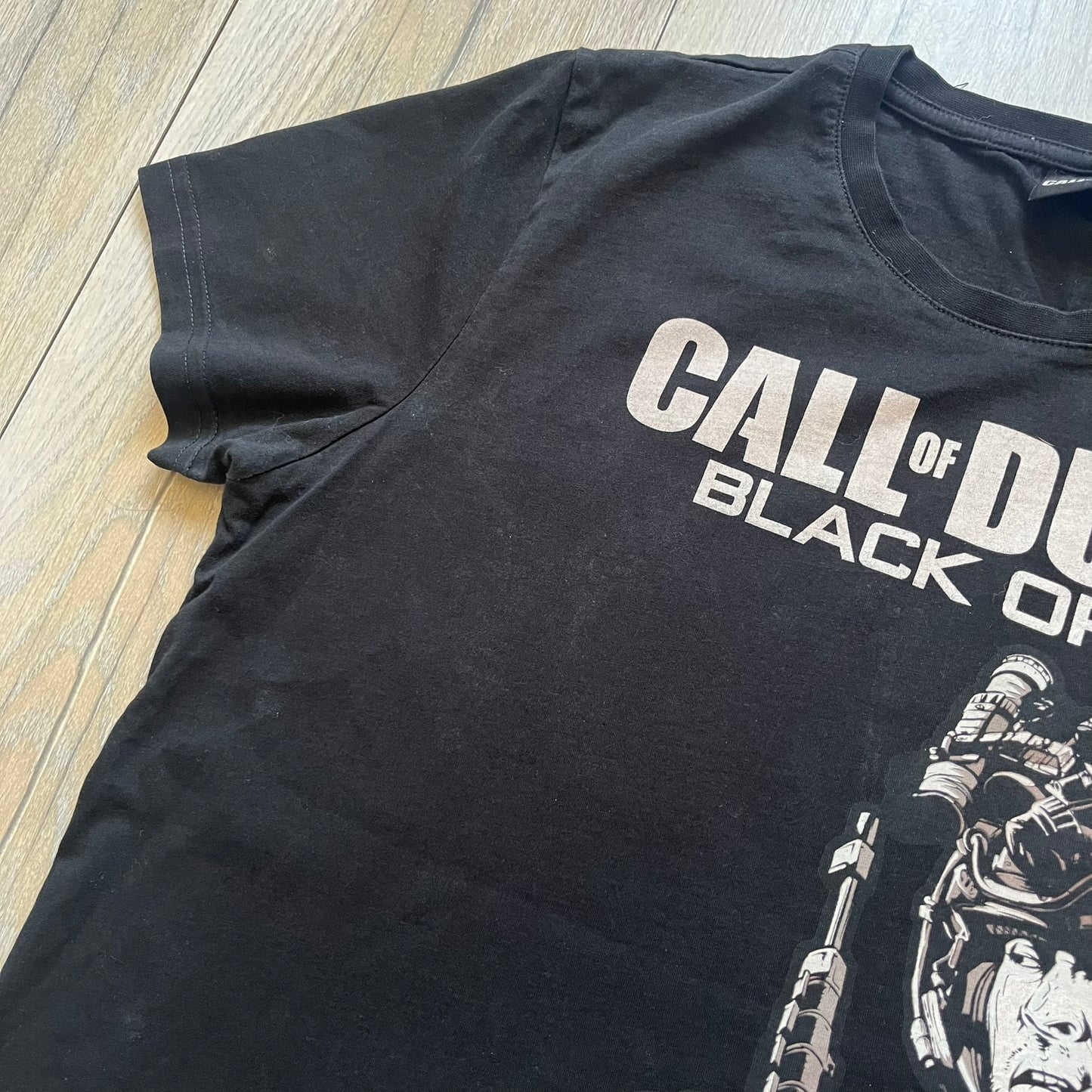Call of Duty: Black Ops 2018 Black Tee is a black promotional T-shirt with a 2018 Activision Publishing licensed neck tag. The front features a large white and grey character graphic in armor holding a weapon, with the “Call of Duty Black Ops” logo printed above. The print spans most of the front from top to bottom.
