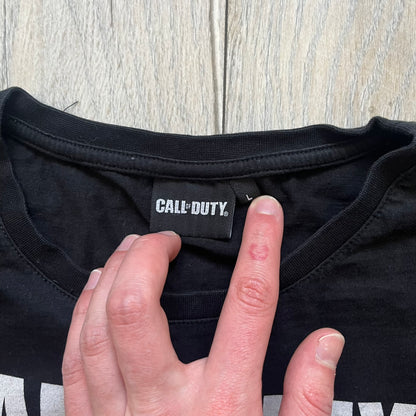 Call of Duty: Black Ops 2018 Black Tee is a black promotional T-shirt with a 2018 Activision Publishing licensed neck tag. The front features a large white and grey character graphic in armor holding a weapon, with the “Call of Duty Black Ops” logo printed above. The print spans most of the front from top to bottom.
