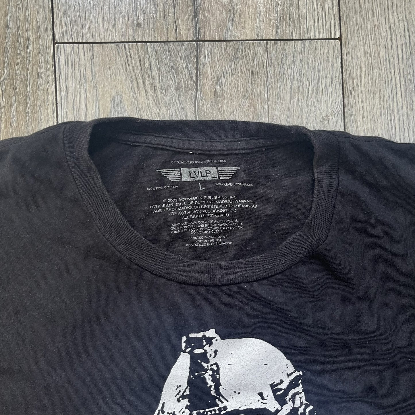 Call of Duty: Modern Warfare 2 2009 Tee is a black T-shirt featuring a large white detailed character with rifle print from the game, under it - green “MW2” text. The sleeve includes an Infinity Ward logo, and the neck tag shows 2009 Activision LVLP branding. Official merchandise from the 2009 Modern Warfare 2 release.