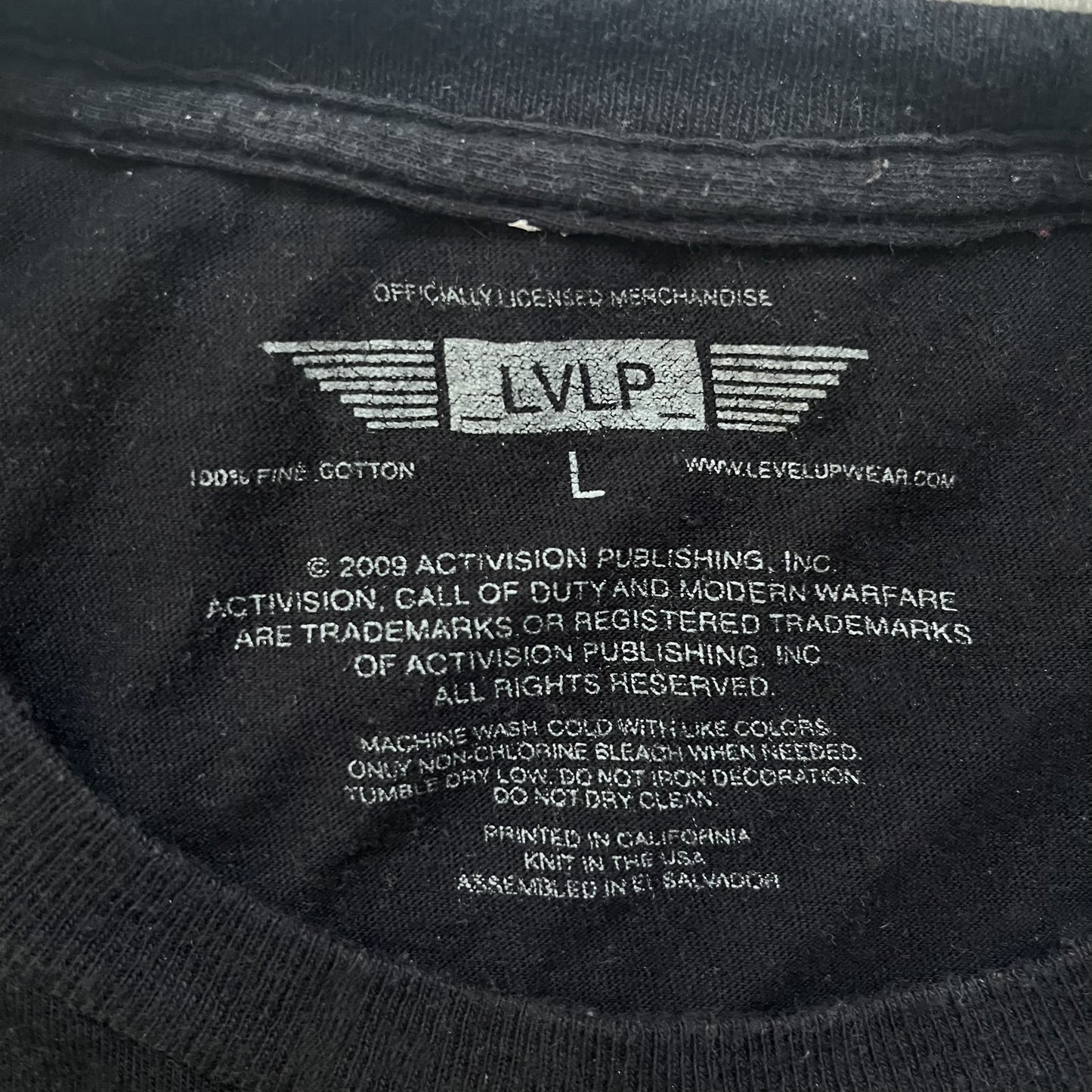 Call of Duty: Modern Warfare 2 2009 Tee is a black T-shirt featuring a large white detailed character with rifle print from the game, under it - green “MW2” text. The sleeve includes an Infinity Ward logo, and the neck tag shows 2009 Activision LVLP branding. Official merchandise from the 2009 Modern Warfare 2 release.