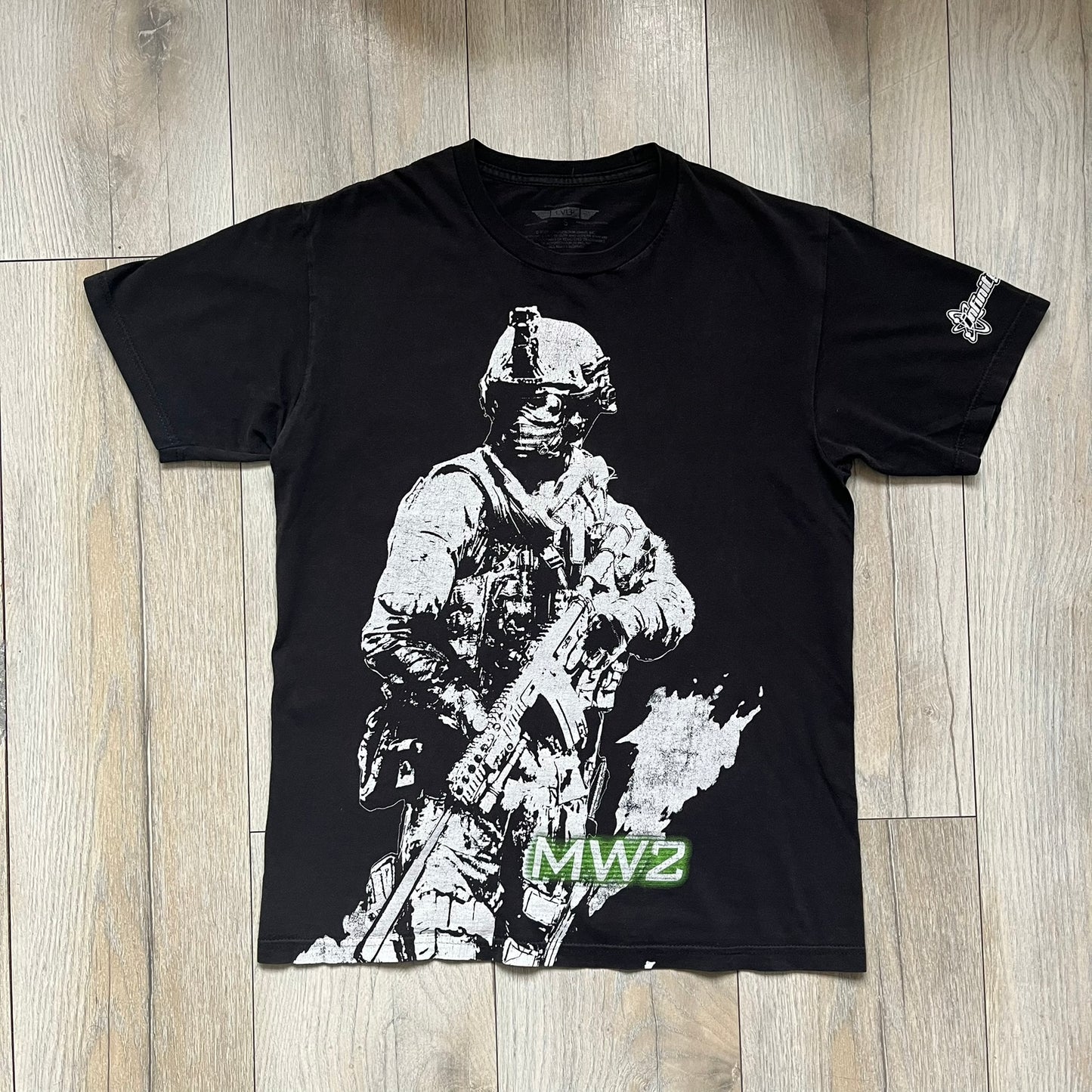 Call of Duty: Modern Warfare 2 2009 Tee is a black T-shirt featuring a large white detailed character print from the game with green “MW2” text below. The sleeve includes an Infinity Ward logo, and the neck tag shows 2009 Activision LVLP branding. Official merchandise from the 2009 Modern Warfare 2 release.