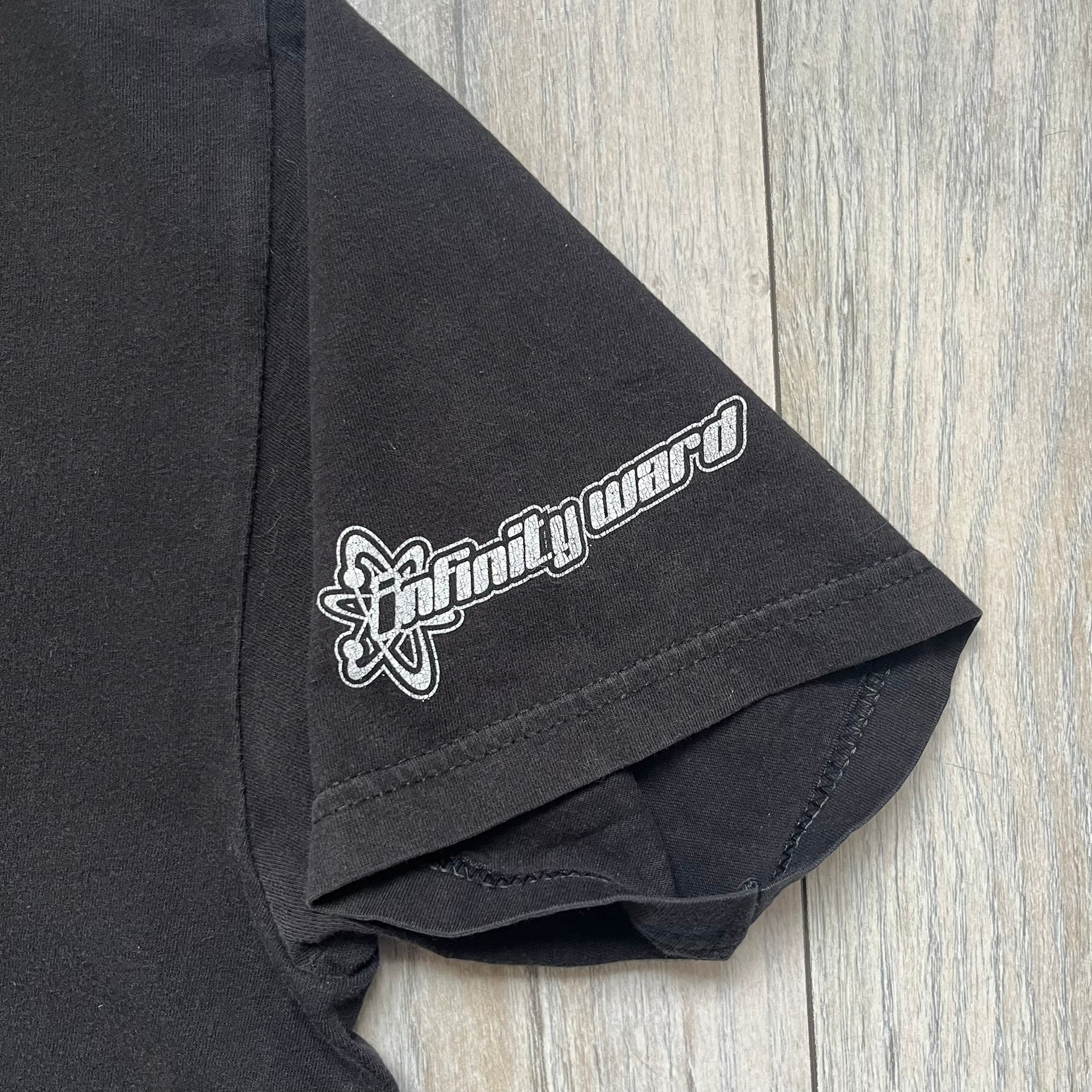 Call of Duty: Modern Warfare 2 2009 Tee is a black T-shirt featuring a large white detailed character print from the game with green “MW2” text below. The sleeve includes an Infinity Ward logo, and the neck tag shows 2009 Activision LVLP branding. Official merchandise from the 2009 Modern Warfare 2 release.