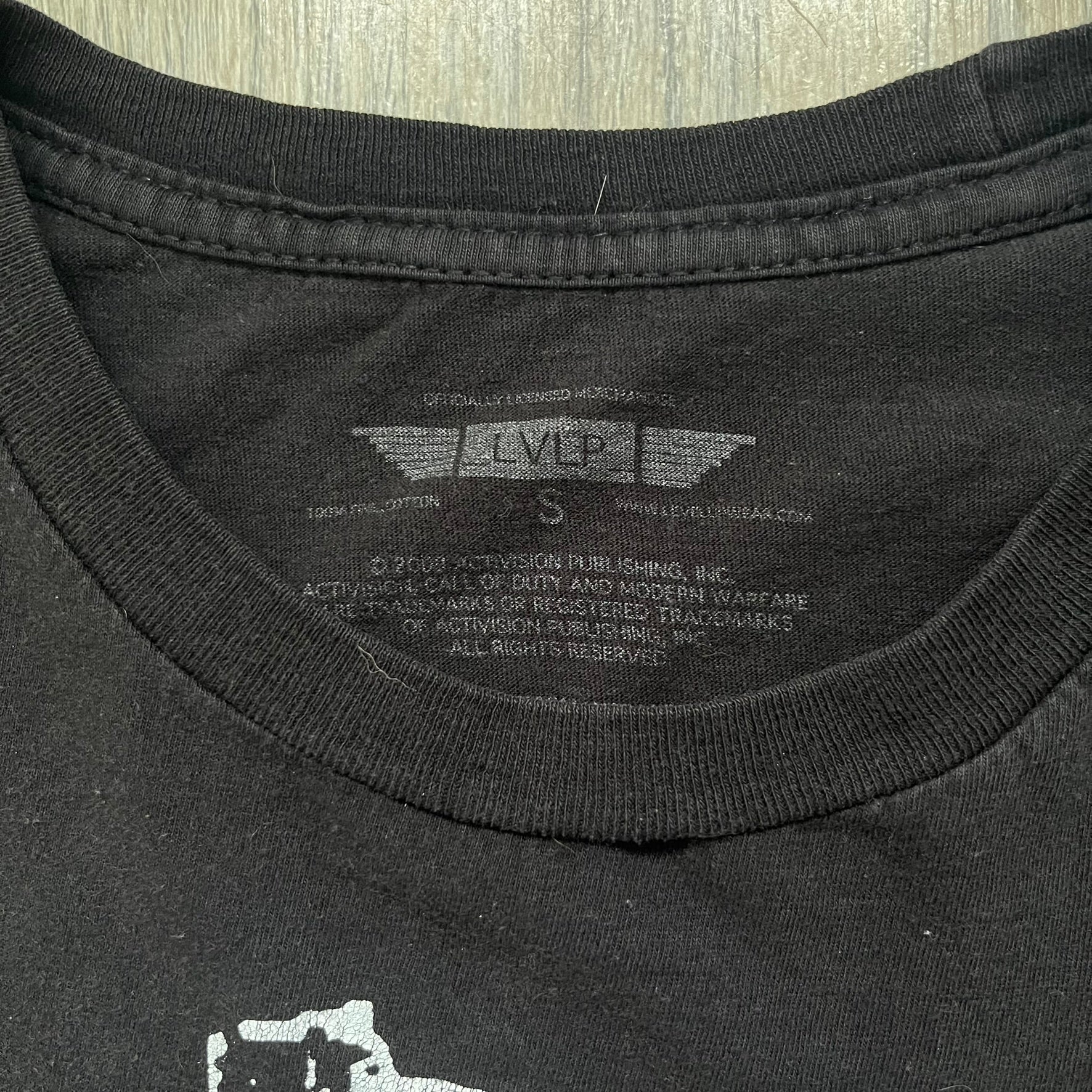 Call of Duty: Modern Warfare 2 2009 Tee is a black T-shirt featuring a large white detailed character print from the game with green “MW2” text below. The sleeve includes an Infinity Ward logo, and the neck tag shows 2009 Activision LVLP branding. Official merchandise from the 2009 Modern Warfare 2 release.