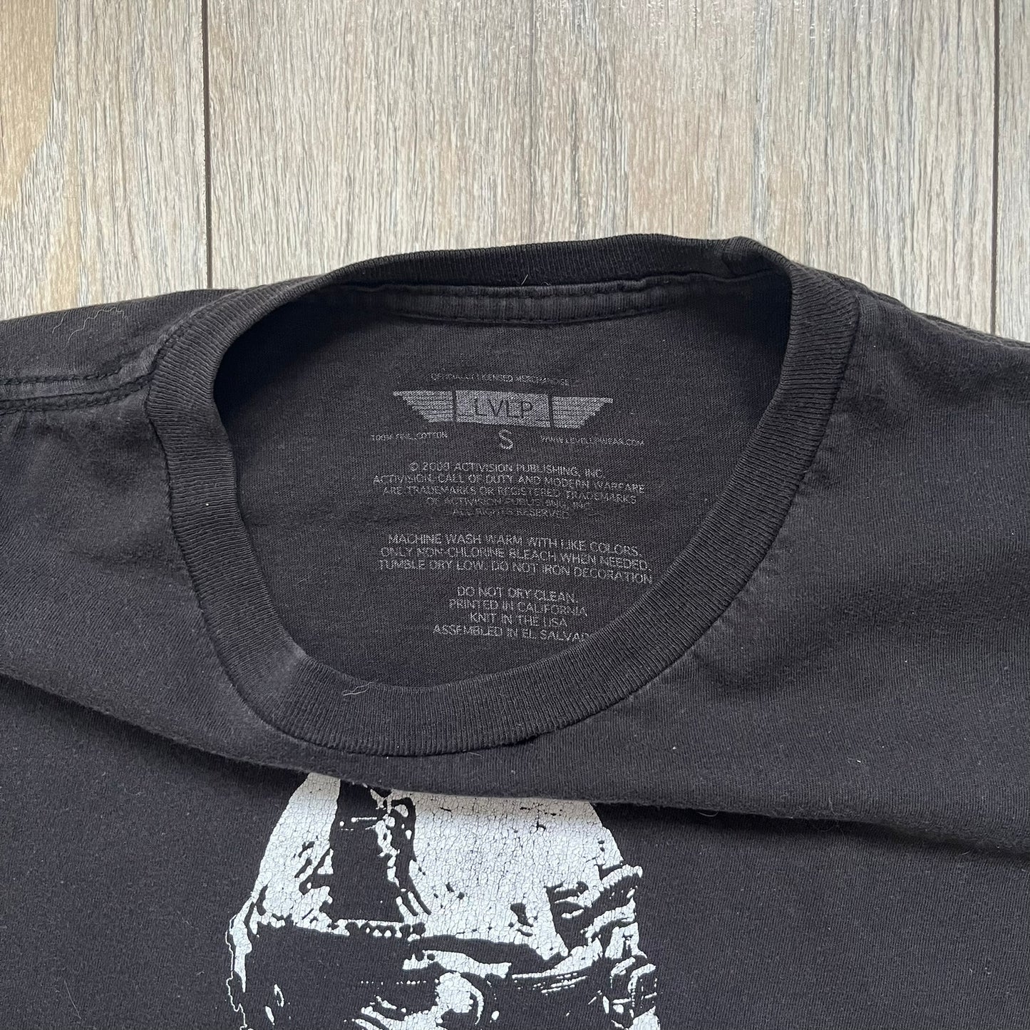 Call of Duty: Modern Warfare 2 2009 Tee is a black T-shirt featuring a large white detailed character print from the game with green “MW2” text below. The sleeve includes an Infinity Ward logo, and the neck tag shows 2009 Activision LVLP branding. Official merchandise from the 2009 Modern Warfare 2 release.