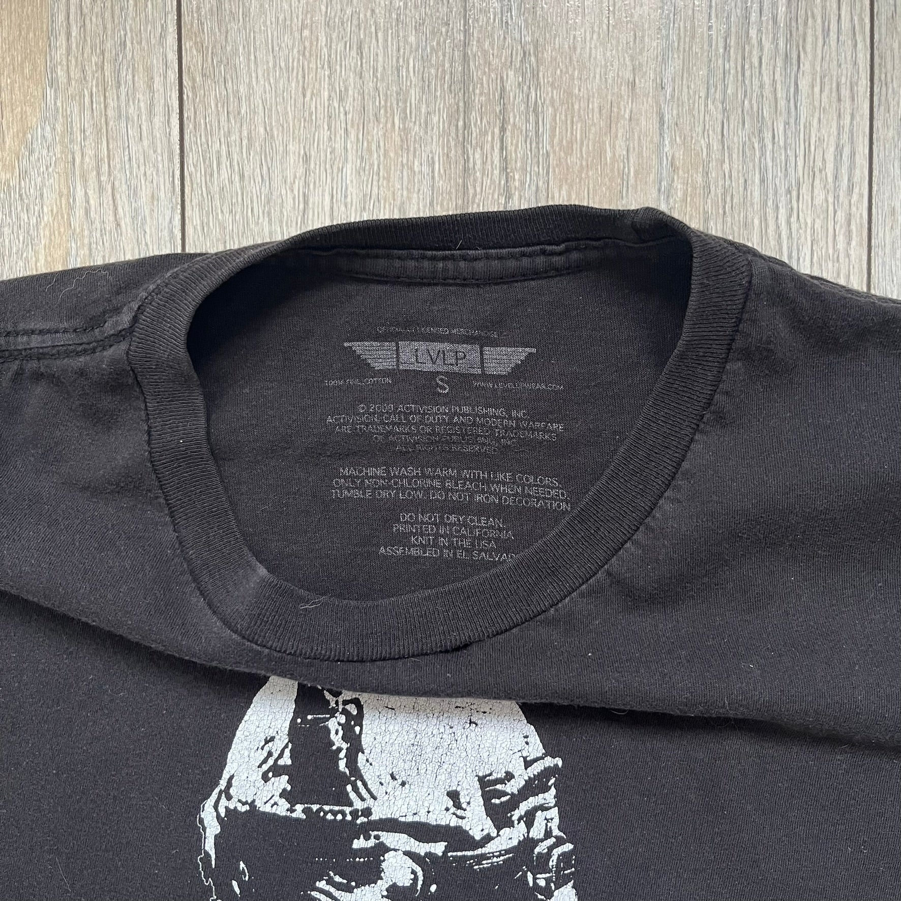 Call of Duty: Modern Warfare 2 2009 Tee is a black T-shirt featuring a large white detailed character print from the game with green “MW2” text below. The sleeve includes an Infinity Ward logo, and the neck tag shows 2009 Activision LVLP branding. Official merchandise from the 2009 Modern Warfare 2 release.