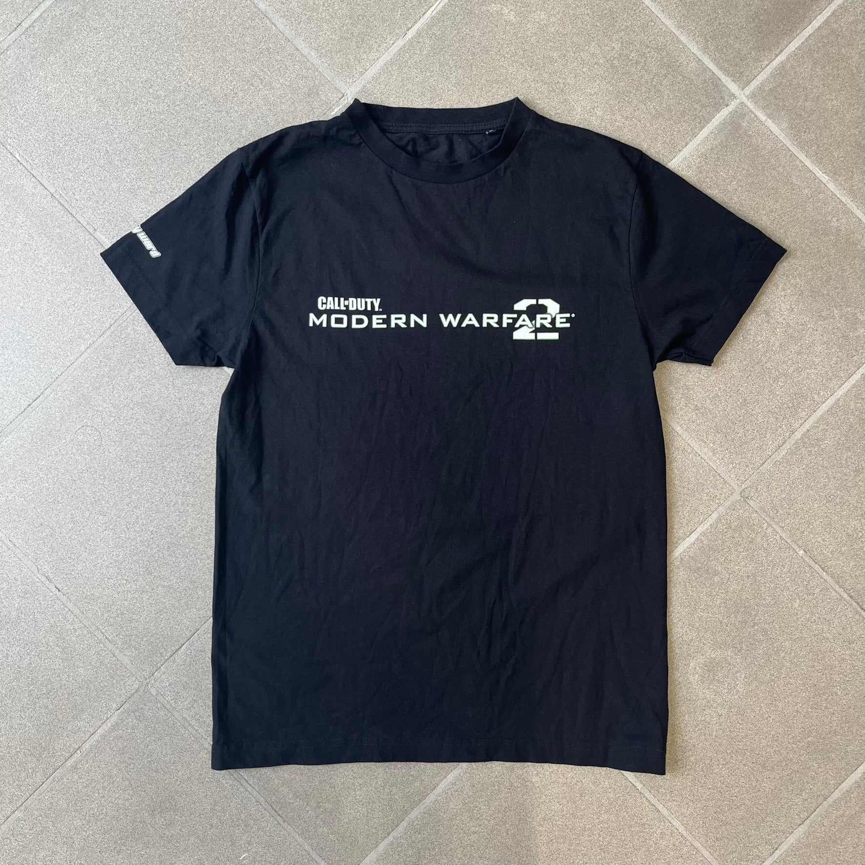 Call of Duty: Modern Warfare 2 2010s Tee features a simple design with the Call of Duty: Modern Warfare 2 logo printed on the front in white and green. The sleeve includes an Infinity Ward logo detail, while the neck has a basic size tag. A later 2010s release inspired by the iconic Modern Warfare 2 branding, not the original promotional tee.
