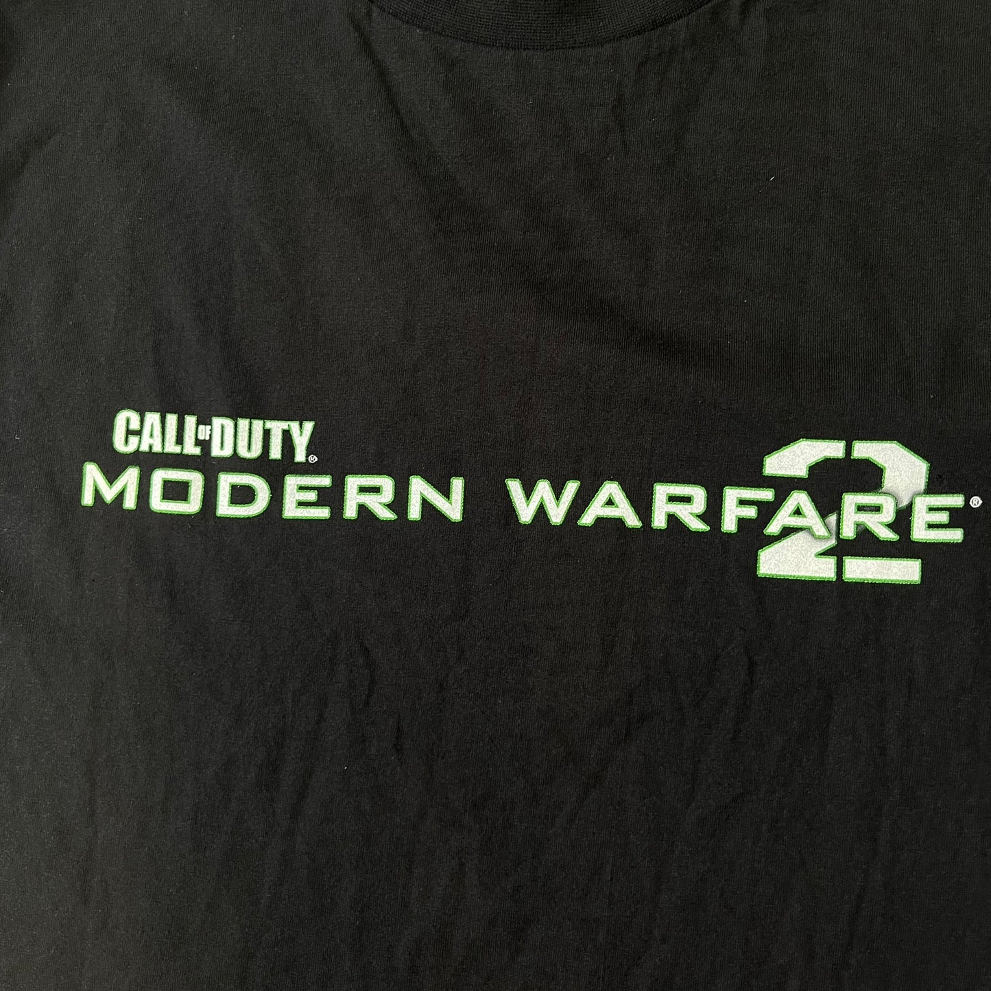 Call of Duty: Modern Warfare 2 2010s Tee features a simple design with the Call of Duty: Modern Warfare 2 logo printed on the front in white and green. The sleeve includes an Infinity Ward logo detail, while the neck has a basic size tag. A later 2010s release inspired by the iconic Modern Warfare 2 branding, not the original promotional tee.
