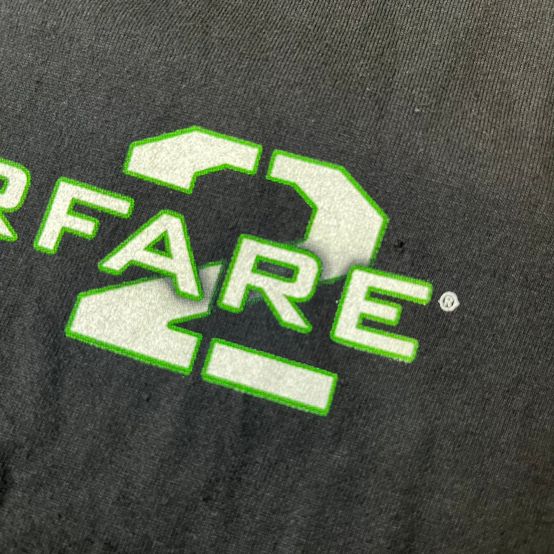 Call of Duty: Modern Warfare 2 2010s Tee features a simple design with the Call of Duty: Modern Warfare 2 logo printed on the front in white and green. The sleeve includes an Infinity Ward logo detail, while the neck has a basic size tag. A later 2010s release inspired by the iconic Modern Warfare 2 branding, not the original promotional tee.
