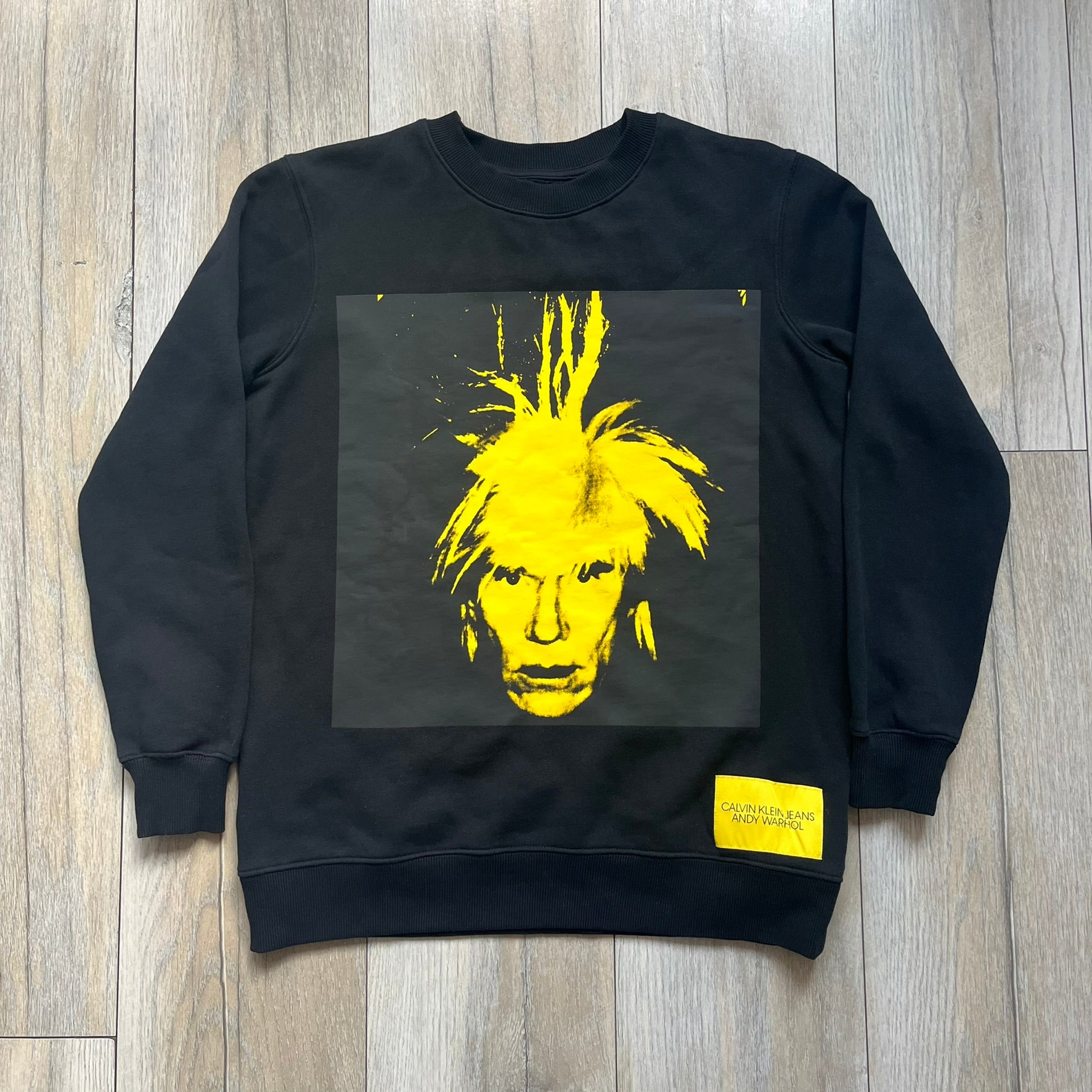 Calvin Klein x Andy Warhol 2018 Self-Portrait Crewneck is a black sweatshirt from the Calvin Klein Jeans x Andy Warhol collaboration. The front features a large yellow self-portrait graphic of Andy Warhol printed on a black background. It includes a yellow Calvin Klein Jeans x Andy Warhol neck tag and a collaboration patch near the bottom hem.
