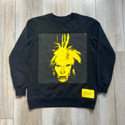 Calvin Klein x Andy Warhol 2018 Self-Portrait Crewneck is a black sweatshirt from the Calvin Klein Jeans x Andy Warhol collaboration. The front features a large yellow self-portrait graphic of Andy Warhol printed on a black background. It includes a yellow Calvin Klein Jeans x Andy Warhol neck tag and a collaboration patch near the bottom hem.

