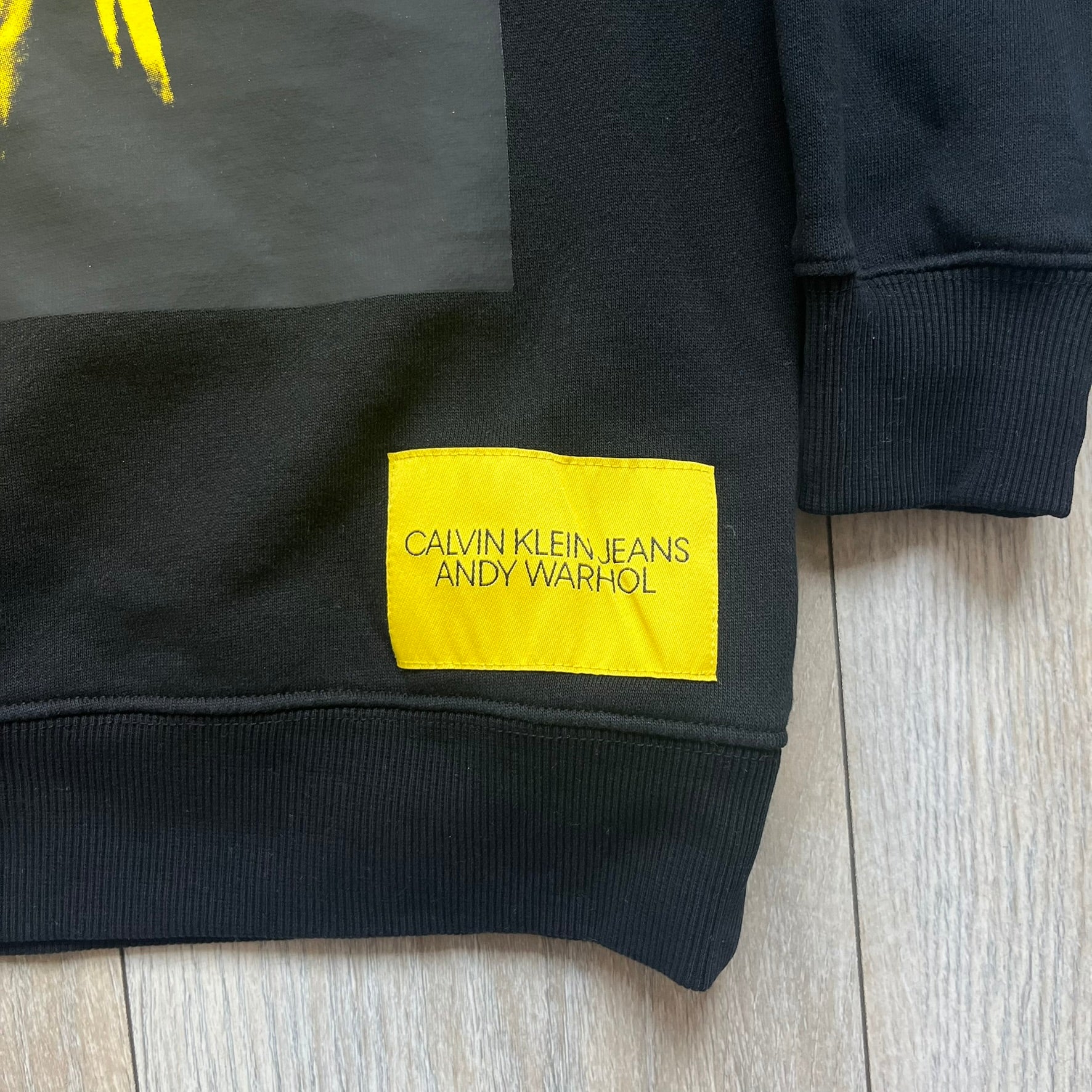 Calvin Klein x Andy Warhol 2018 Self-Portrait Crewneck is a black sweatshirt from the Calvin Klein Jeans x Andy Warhol collaboration. The front features a large yellow self-portrait graphic of Andy Warhol printed on a black background. It includes a yellow Calvin Klein Jeans x Andy Warhol neck tag and a collaboration patch near the bottom hem.
