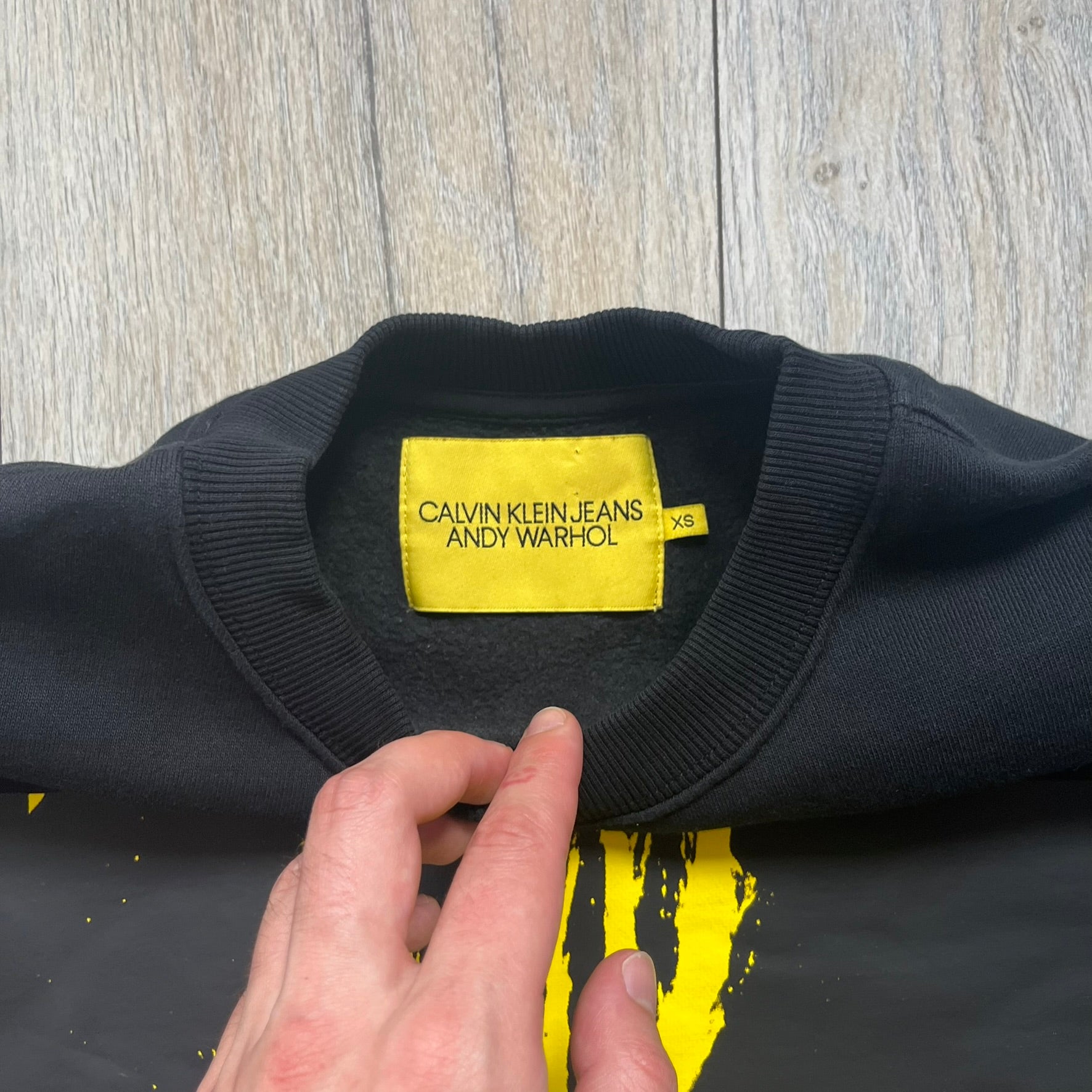 Calvin Klein x Andy Warhol 2018 Self-Portrait Crewneck is a black sweatshirt from the Calvin Klein Jeans x Andy Warhol collaboration. The front features a large yellow self-portrait graphic of Andy Warhol printed on a black background. It includes a yellow Calvin Klein Jeans x Andy Warhol neck tag and a collaboration patch near the bottom hem.
