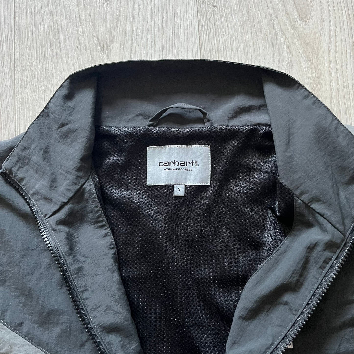 Carhartt WIP Gray Field Windbreaker featuring a dynamic two-tone design split diagonally across the body in light and dark gray. Crafted from durable nylon and polyester layers, it includes a small white Carhartt logo on the chest and large diagonal Carhartt embroidery on the back.