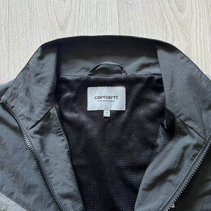 Carhartt WIP Gray Field Windbreaker featuring a dynamic two-tone design split diagonally across the body in light and dark gray. Crafted from durable nylon and polyester layers, it includes a small white Carhartt logo on the chest and large diagonal Carhartt embroidery on the back.