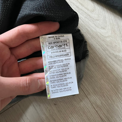 Carhartt WIP Gray Field Windbreaker featuring a dynamic two-tone design split diagonally across the body in light and dark gray. Crafted from durable nylon and polyester layers, it includes a small white Carhartt logo on the chest and large diagonal Carhartt embroidery on the back.