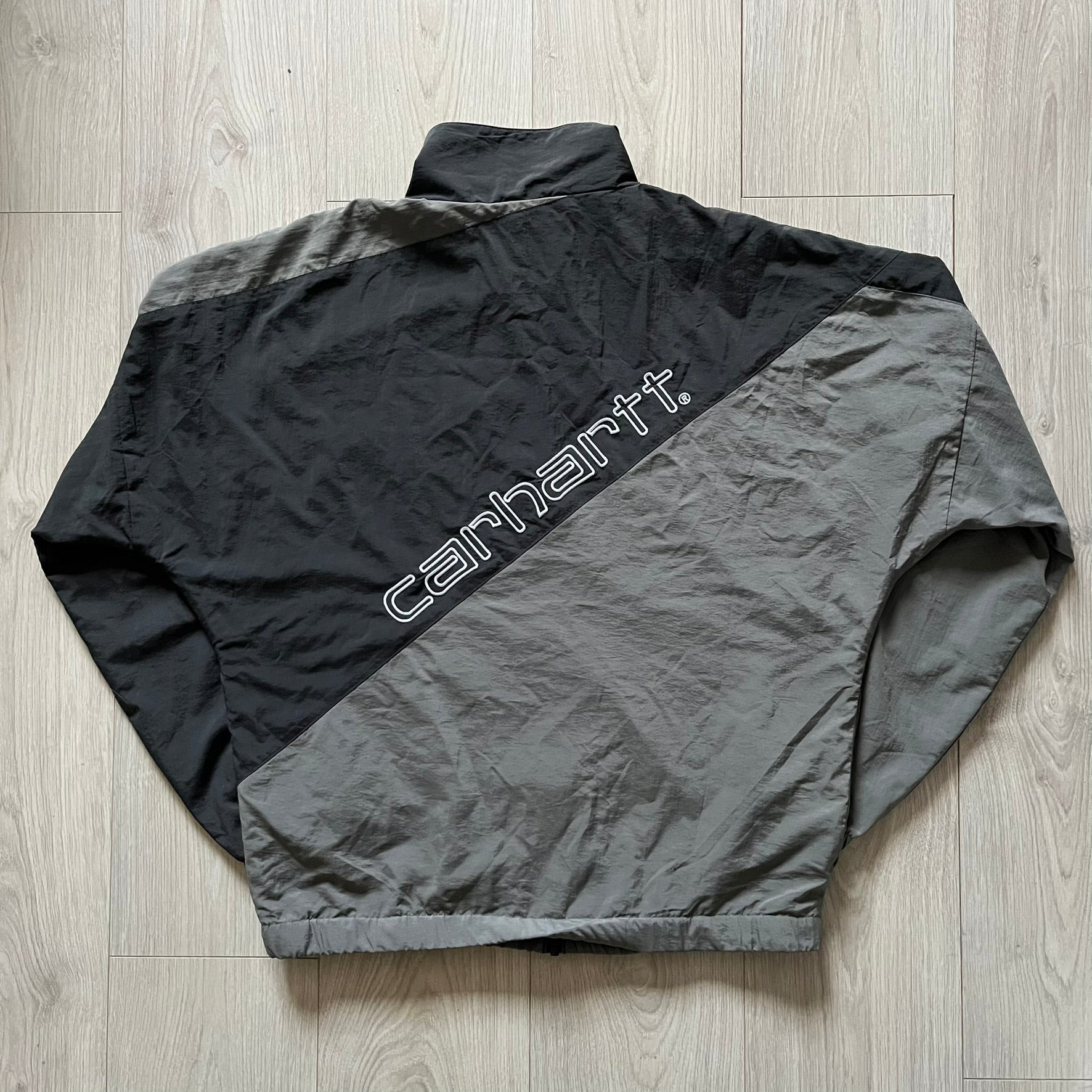 Carhartt WIP Gray Field Windbreaker featuring a dynamic two-tone design split diagonally across the body in light and dark gray. Crafted from durable nylon and polyester layers, it includes a small white Carhartt logo on the chest and large diagonal Carhartt embroidery on the back.