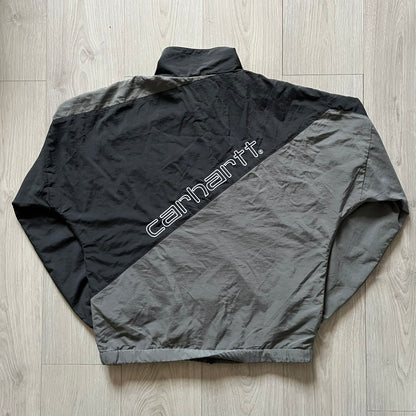 Carhartt WIP Gray Field Windbreaker featuring a dynamic two-tone design split diagonally across the body in light and dark gray. Crafted from durable nylon and polyester layers, it includes a small white Carhartt logo on the chest and large diagonal Carhartt embroidery on the back.