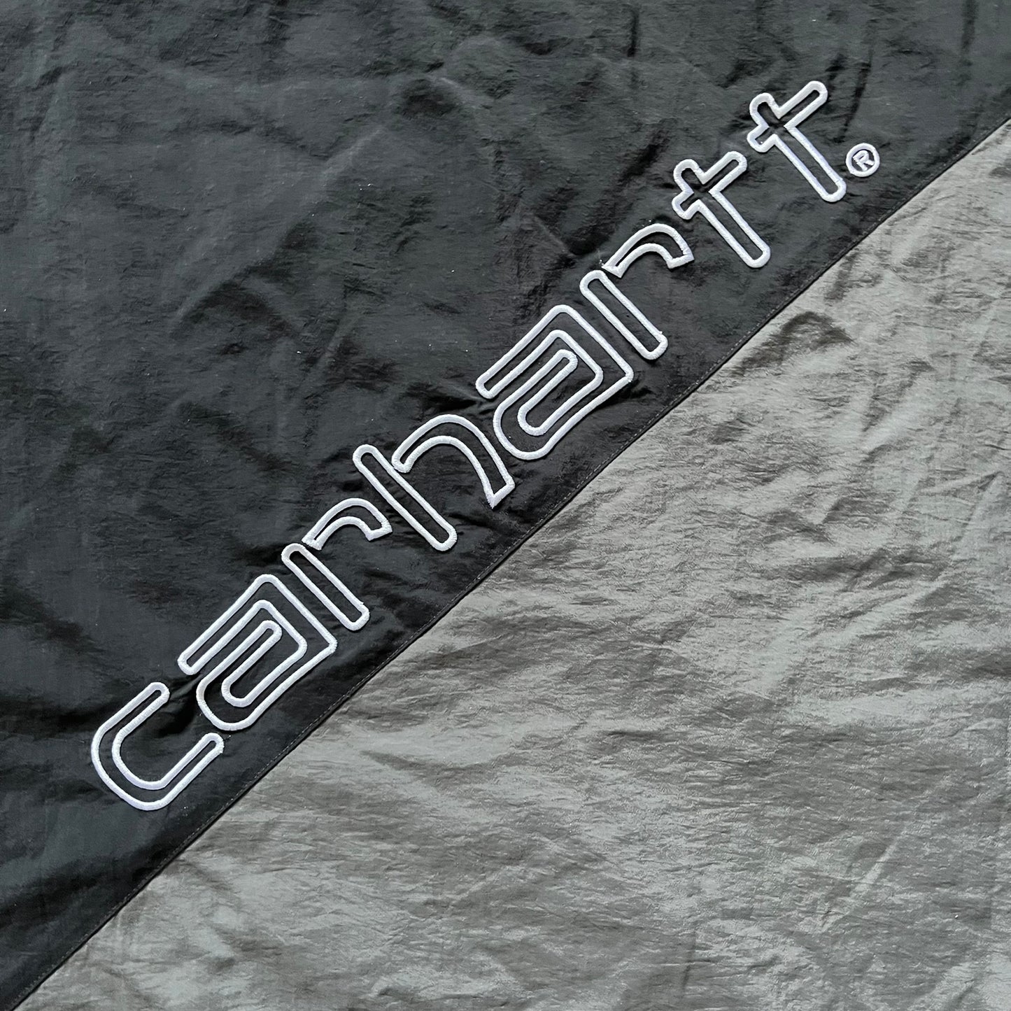 Carhartt WIP Gray Field Windbreaker featuring a dynamic two-tone design split diagonally across the body in light and dark gray. Crafted from durable nylon and polyester layers, it includes a small white Carhartt logo on the chest and large diagonal Carhartt embroidery on the back.