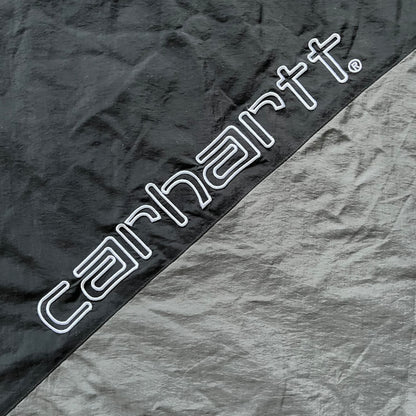 Carhartt WIP Gray Field Windbreaker featuring a dynamic two-tone design split diagonally across the body in light and dark gray. Crafted from durable nylon and polyester layers, it includes a small white Carhartt logo on the chest and large diagonal Carhartt embroidery on the back.
