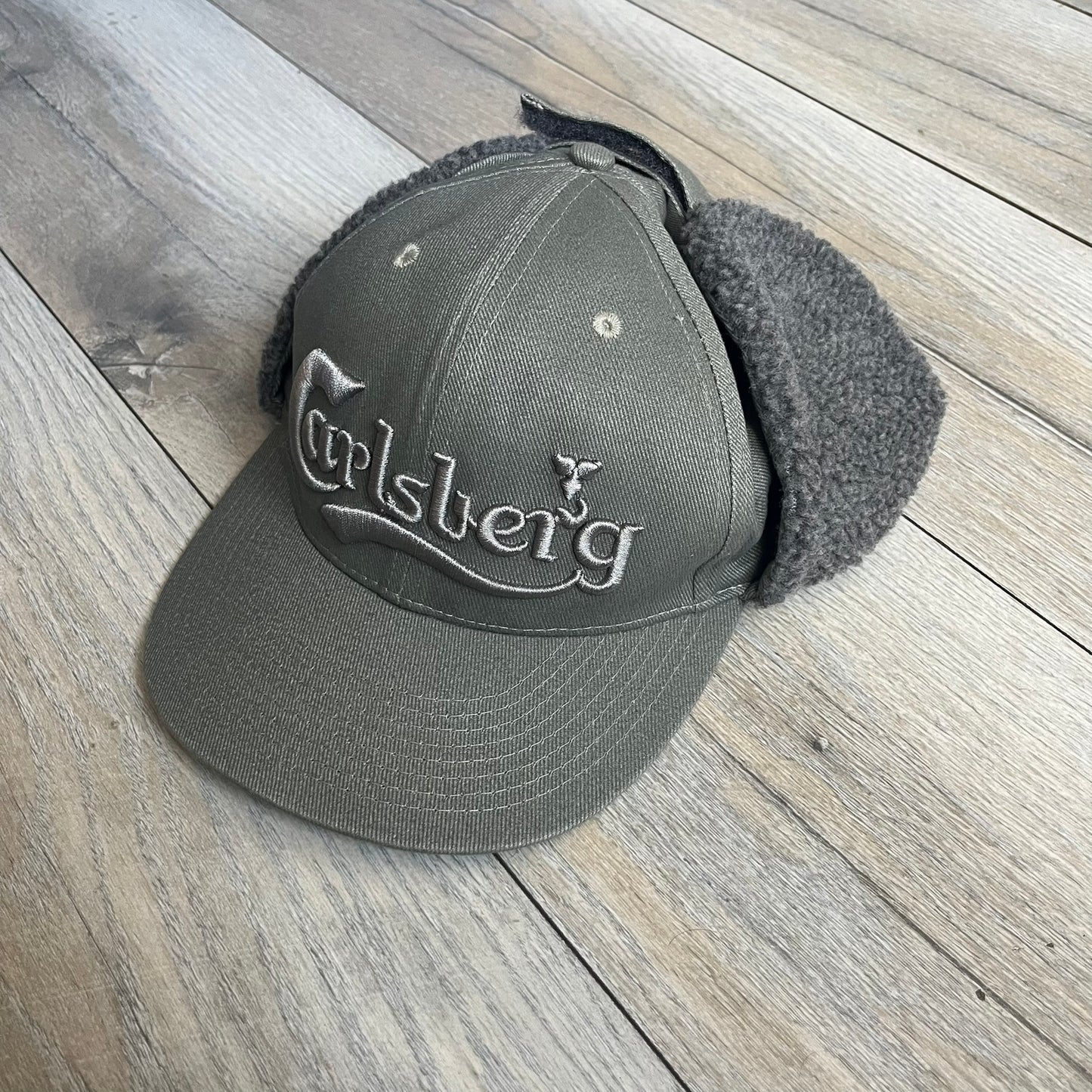 Carlsberg Gray Earflaps Hat is a gray snapback cap featuring a large embroidered Carlsberg logo across the front. The back includes a small leather logo patch, while the sides are fitted with soft earflaps that can be secured under the chin using Velcro straps.
