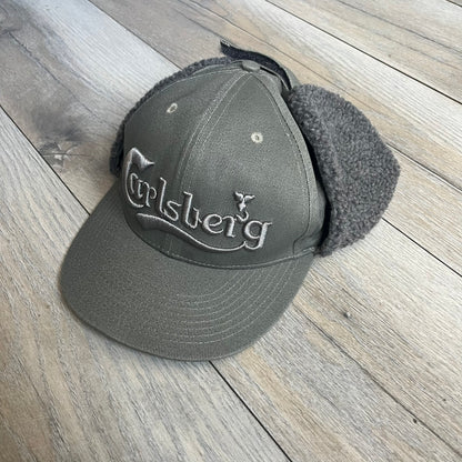 Carlsberg Gray Earflaps Hat is a gray snapback cap featuring a large embroidered Carlsberg logo across the front. The back includes a small leather logo patch, while the sides are fitted with soft earflaps that can be secured under the chin using Velcro straps.

