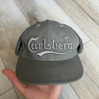 Carlsberg Gray Earflaps Hat is a gray snapback cap featuring a large embroidered Carlsberg logo across the front. The back includes a small leather logo patch, while the sides are fitted with soft earflaps that can be secured under the chin using Velcro straps.
