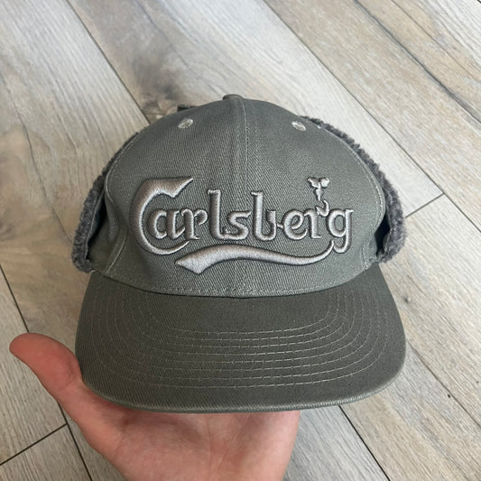 Carlsberg Gray Earflaps Hat is a gray snapback cap featuring a large embroidered Carlsberg logo across the front. The back includes a small leather logo patch, while the sides are fitted with soft earflaps that can be secured under the chin using Velcro straps.
