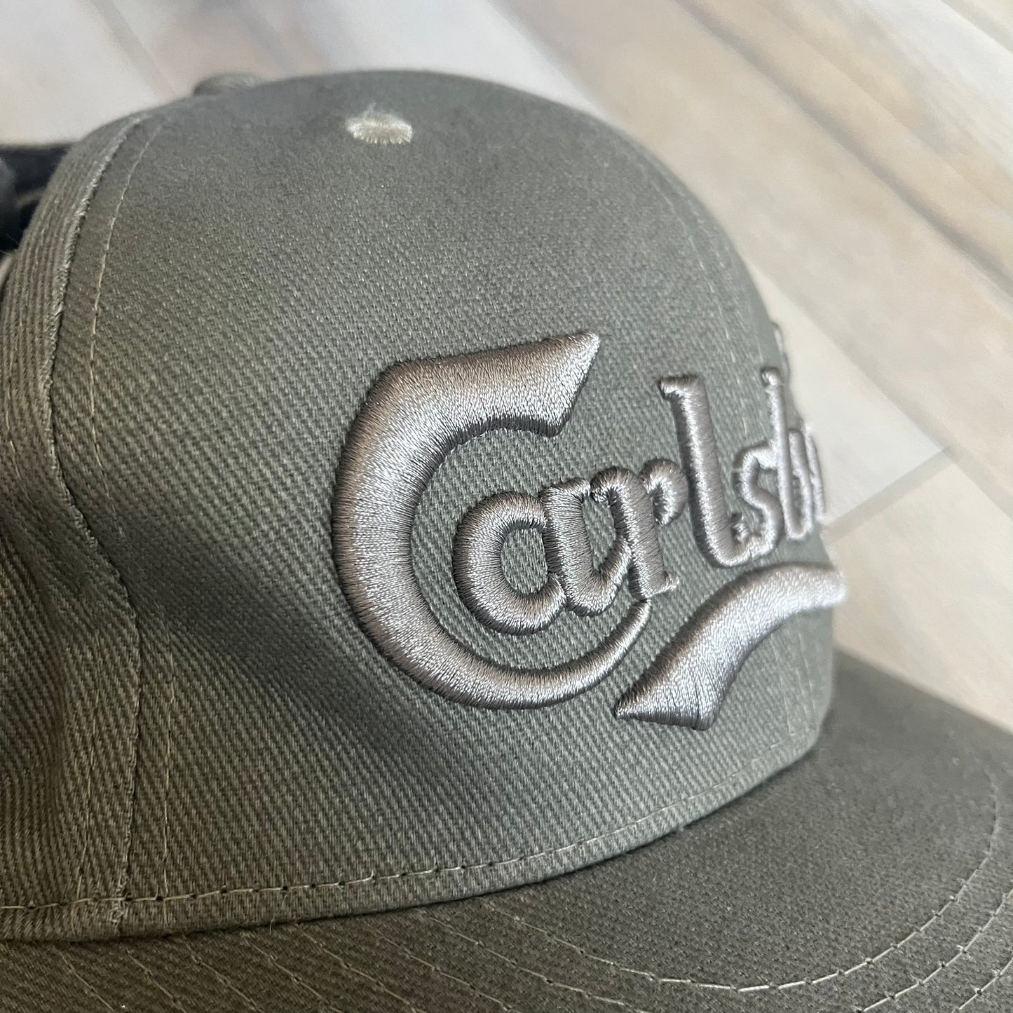 Carlsberg Gray Earflaps Hat is a gray snapback cap featuring a large embroidered Carlsberg logo across the front. The back includes a small leather logo patch, while the sides are fitted with soft earflaps that can be secured under the chin using Velcro straps.
