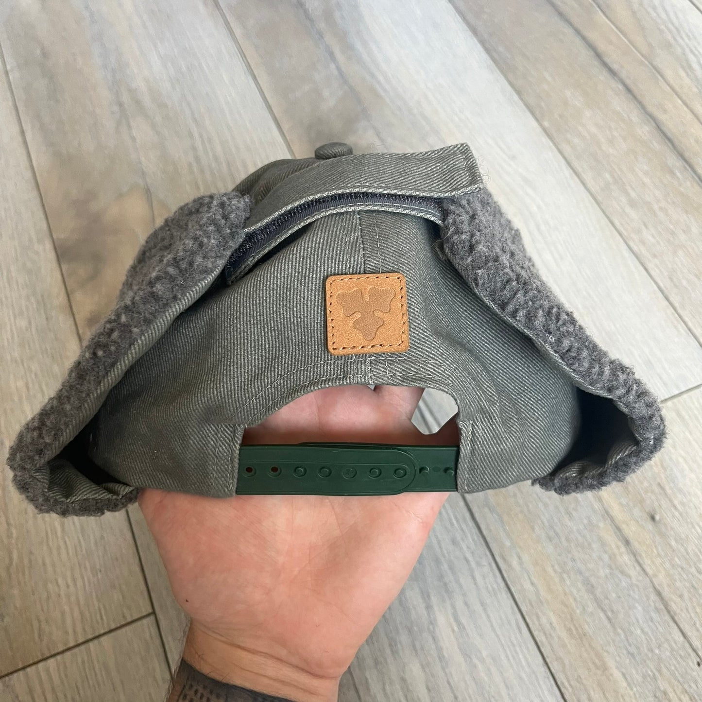 Carlsberg Gray Earflaps Hat is a gray snapback cap featuring a large embroidered Carlsberg logo across the front. The back includes a small leather logo patch, while the sides are fitted with soft earflaps that can be secured under the chin using Velcro straps.
