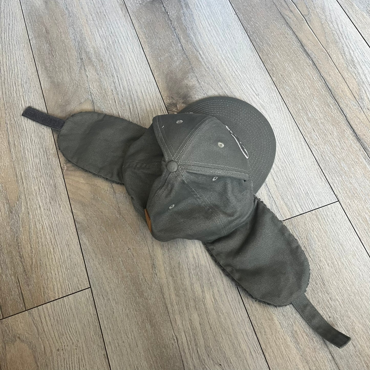 Carlsberg Gray Earflaps Hat is a gray snapback cap featuring a large embroidered Carlsberg logo across the front. The back includes a small leather logo patch, while the sides are fitted with soft earflaps that can be secured under the chin using Velcro straps.
