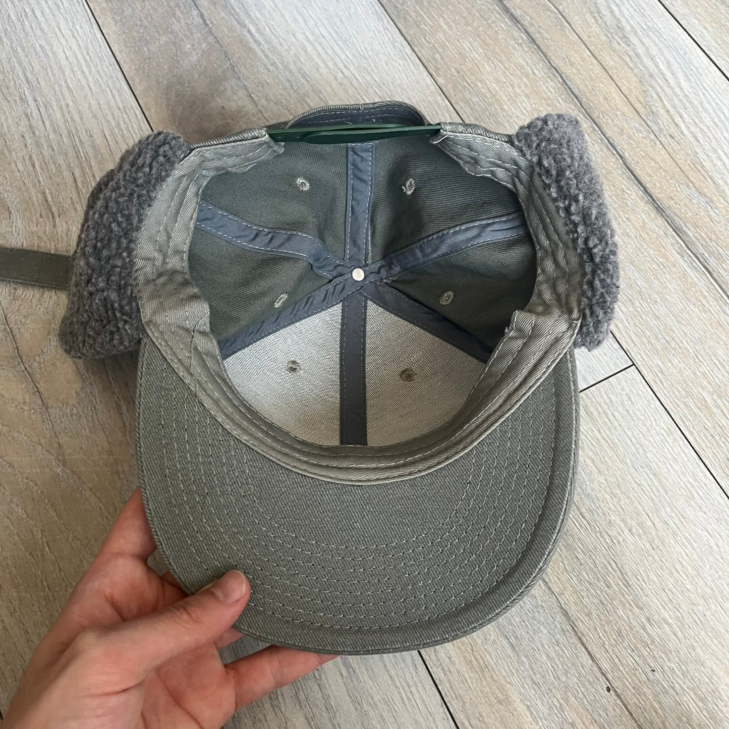Carlsberg Gray Earflaps Hat is a gray snapback cap featuring a large embroidered Carlsberg logo across the front. The back includes a small leather logo patch, while the sides are fitted with soft earflaps that can be secured under the chin using Velcro straps.
