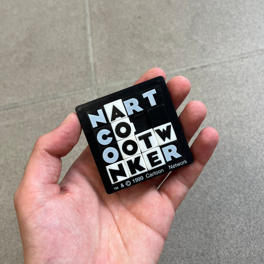 Cartoon Network 1999 Logo Puzzle is a handheld sliding tile puzzle featuring the classic Cartoon Network logo. The movable square tiles each display a single black or white letter, allowing the logo to be rearranged by sliding the pieces into place. Official 1999 licensing is printed on the bottom of the puzzle frame.
