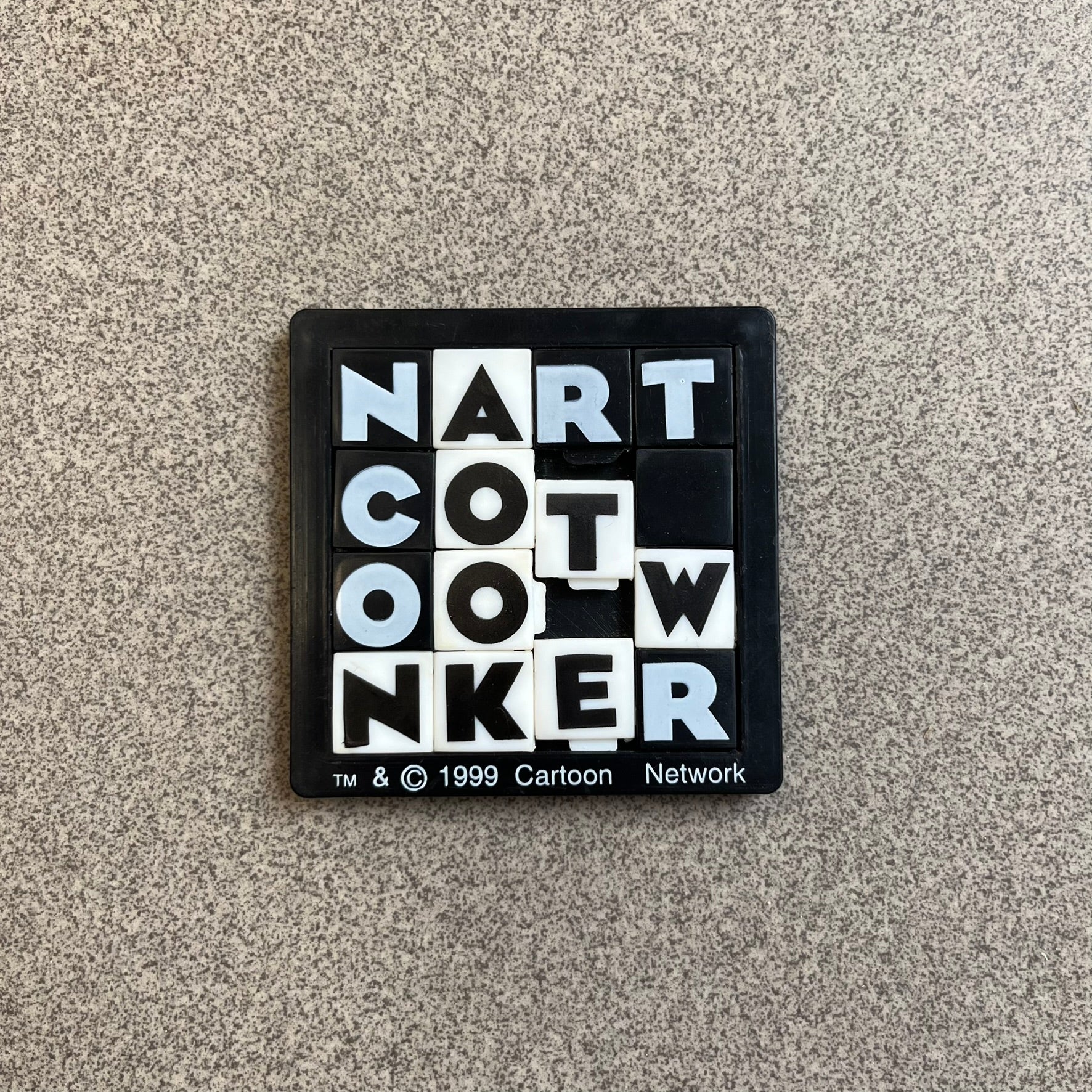Cartoon Network 1999 Logo Puzzle is a handheld sliding tile puzzle featuring the classic Cartoon Network logo. The movable square tiles each display a single black or white letter, allowing the logo to be rearranged by sliding the pieces into place. Official 1999 licensing is printed on the bottom of the puzzle frame.