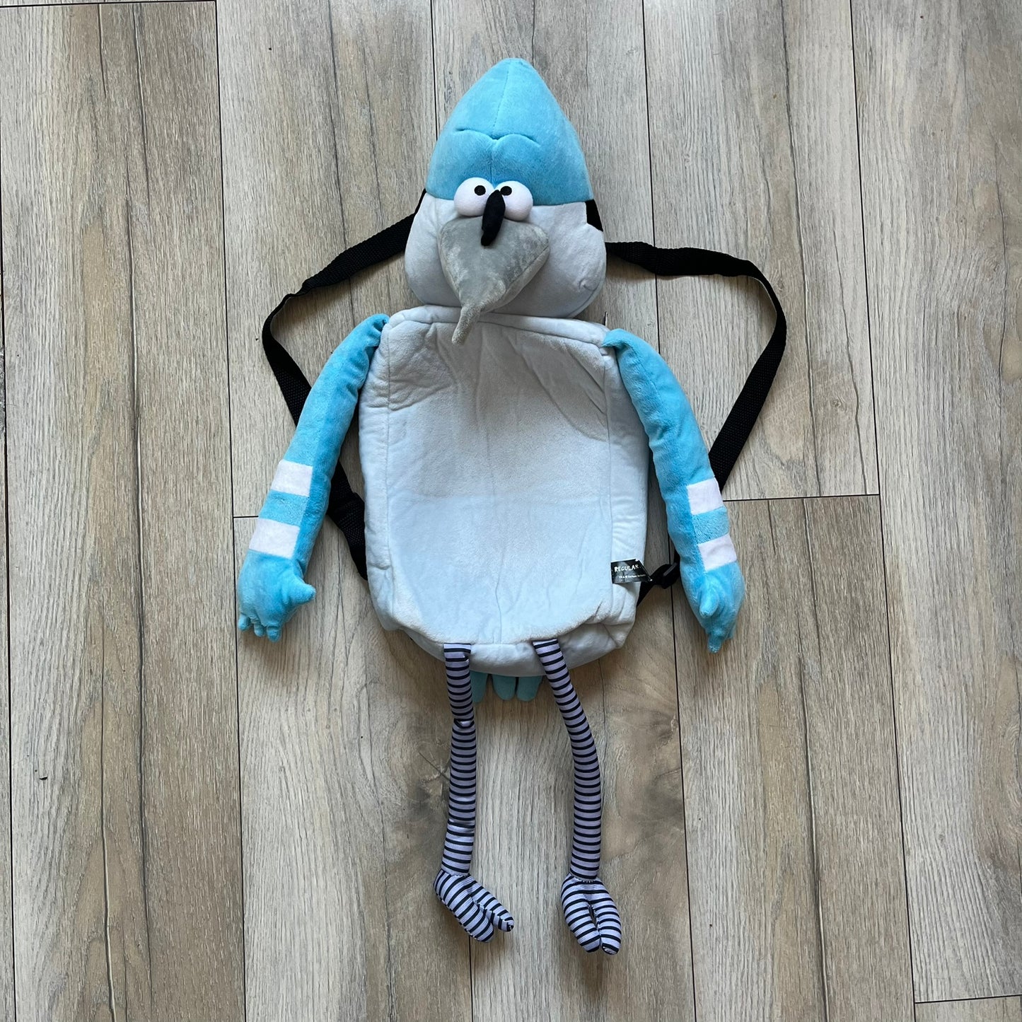 Cartoon Network Regular Show Mordecai 2016 Backpack is a full-body backpack featuring Mordecai with movable arms and legs. The straps attach from top to bottom, and the design shows his blue and light blue coloring, black head markings, striped wings, and tail feathers. The backpack replicates Mordecai’s animated appearance, including his crested head feathers and grey striped legs.