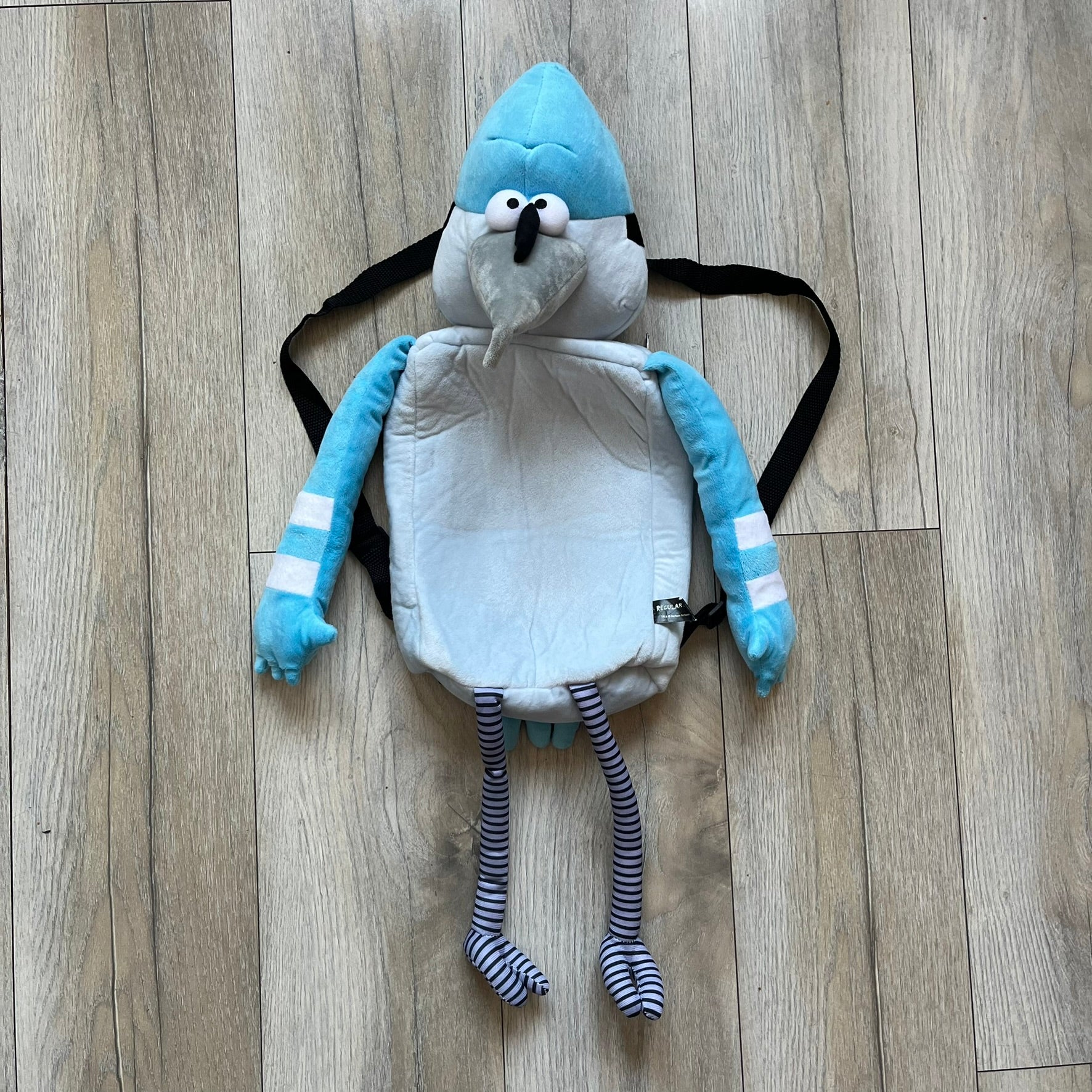 Cartoon Network Regular Show Mordecai 2016 Backpack is a full-body backpack featuring Mordecai with movable arms and legs. The straps attach from top to bottom, and the design shows his blue and light blue coloring, black head markings, striped wings, and tail feathers. The backpack replicates Mordecai’s animated appearance, including his crested head feathers and grey striped legs.