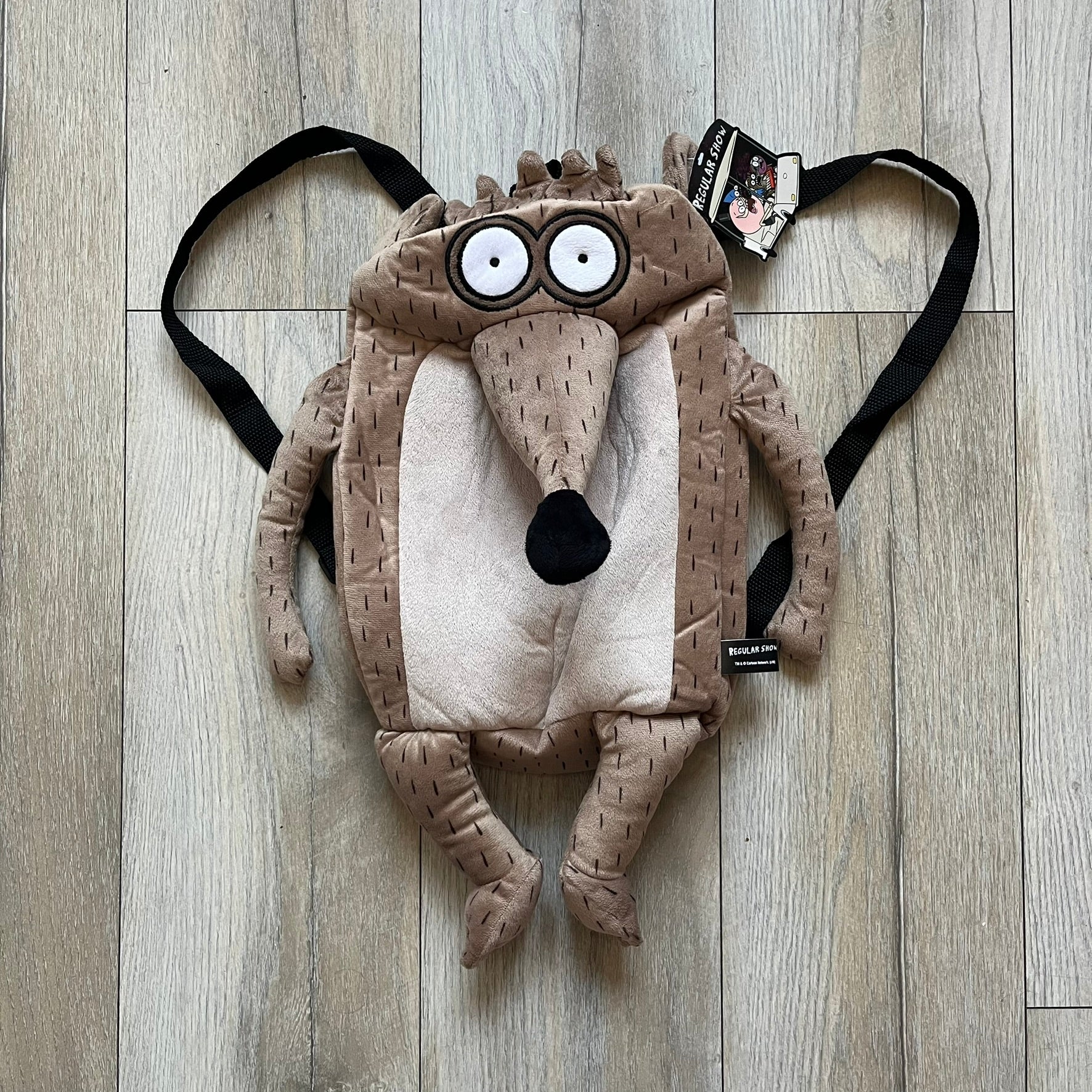 Cartoon Network Regular Show Rigby 2016 Backpack is a character-shaped backpack designed as Rigby from the animated series Regular Show. The bag features a full plush body construction with movable arms, legs, and mouth, while the eyes and mouth are finished with embroidered detailing. Adjustable straps are attached from the top to the lower back of the character, allowing it to be worn as a standard backpack. The design follows Rigby’s appearance with brown fur tones, dark raccoon-style eye mask markings, 