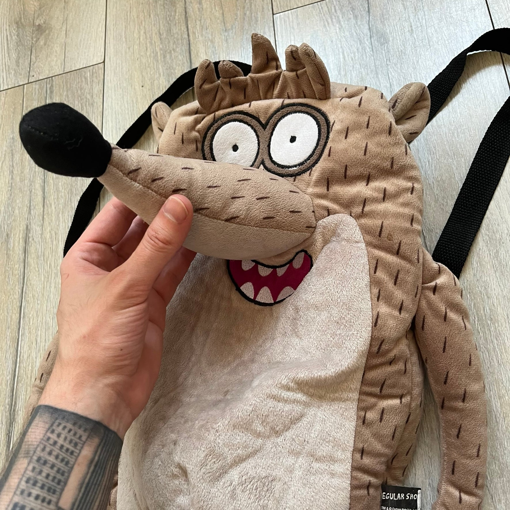 Cartoon Network Regular Show Rigby 2016 Backpack is a character-shaped backpack designed as Rigby from the animated series Regular Show. The bag features a full plush body construction with movable arms, legs, and mouth, while the eyes and mouth are finished with embroidered detailing. Adjustable straps are attached from the top to the lower back of the character, allowing it to be worn as a standard backpack. The design follows Rigby’s appearance with brown fur tones, dark raccoon-style eye mask markings, 