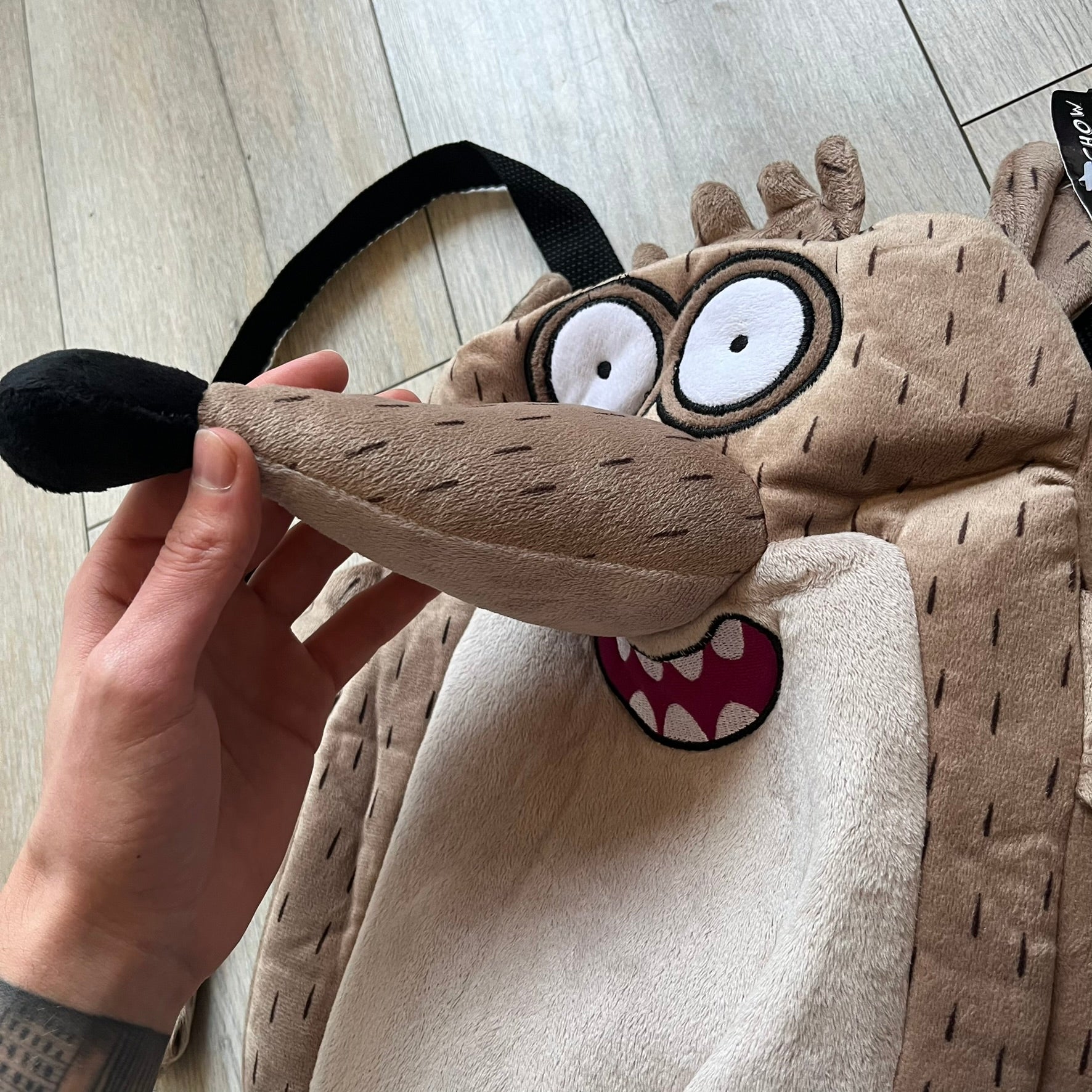 Cartoon Network Regular Show Rigby 2016 Backpack is a character-shaped backpack designed as Rigby from the animated series Regular Show. The bag features a full plush body construction with movable arms, legs, and mouth, while the eyes and mouth are finished with embroidered detailing. Adjustable straps are attached from the top to the lower back of the character, allowing it to be worn as a standard backpack. The design follows Rigby’s appearance with brown fur tones, dark raccoon-style eye mask markings, 