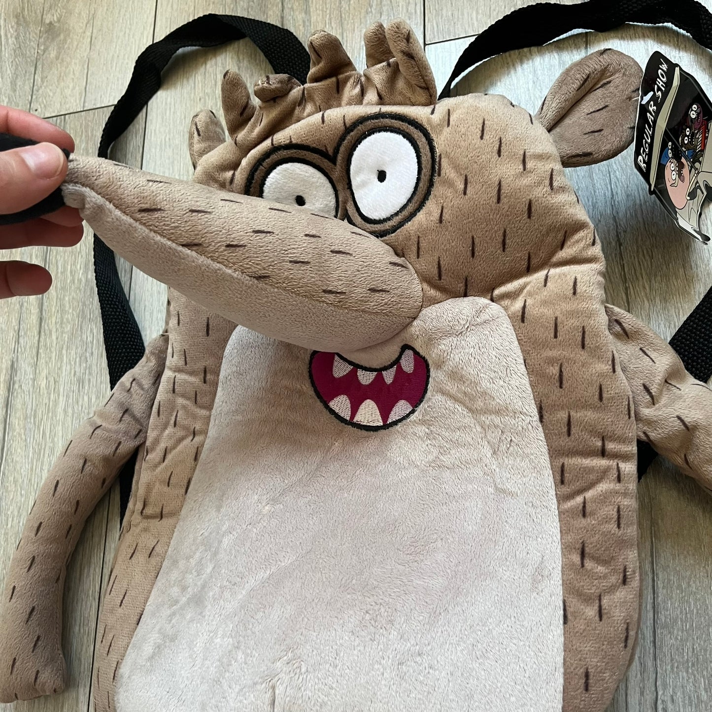 Cartoon Network Regular Show Rigby 2016 Backpack is a character-shaped backpack designed as Rigby from the animated series Regular Show. The bag features a full plush body construction with movable arms, legs, and mouth, while the eyes and mouth are finished with embroidered detailing. Adjustable straps are attached from the top to the lower back of the character, allowing it to be worn as a standard backpack. The design follows Rigby’s appearance with brown fur tones, dark raccoon-style eye mask markings, 