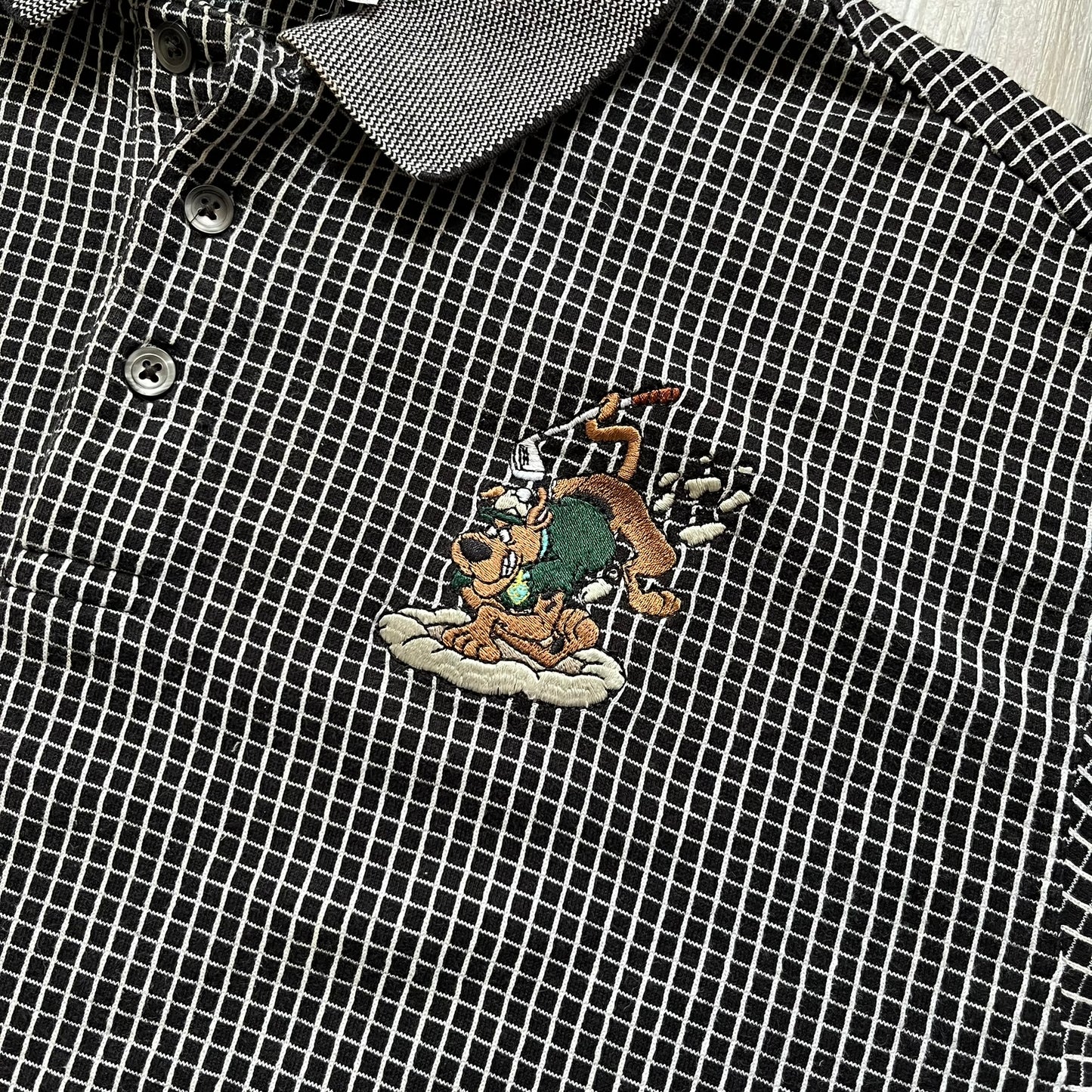 Cartoon Network Scooby-Doo 1999 Golf Polo is a black polo shirt featuring a grid pattern of thin white lines forming small squares across the fabric. The left chest displays an embroidered graphic of Scooby-Doo in a golf outfit digging in sand. The neck tag includes Warner Bros. and Cartoon Network 1999 licensing.