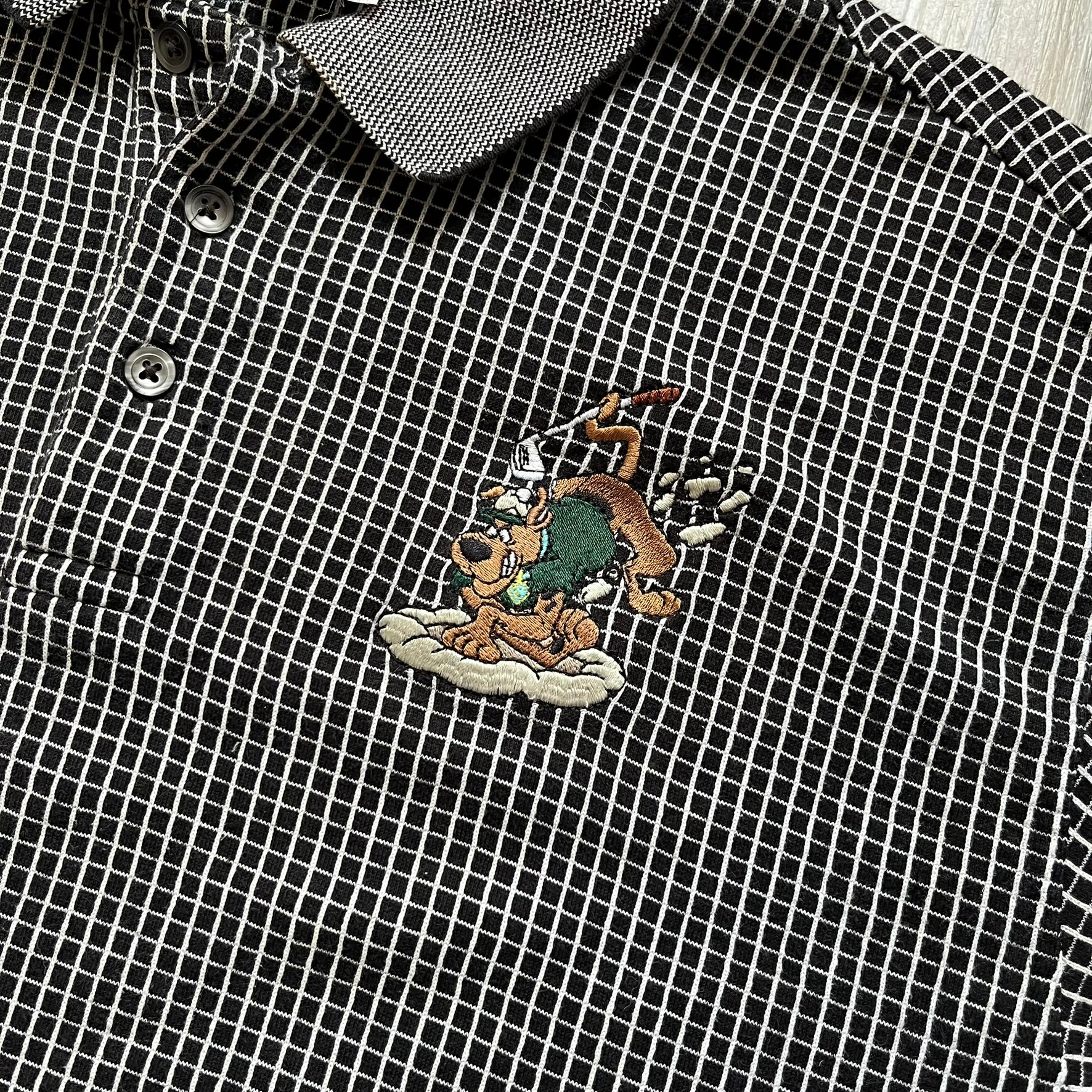 Cartoon Network Scooby-Doo 1999 Golf Polo is a black polo shirt featuring a grid pattern of thin white lines forming small squares across the fabric. The left chest displays an embroidered graphic of Scooby-Doo in a golf outfit digging in sand. The neck tag includes Warner Bros. and Cartoon Network 1999 licensing.