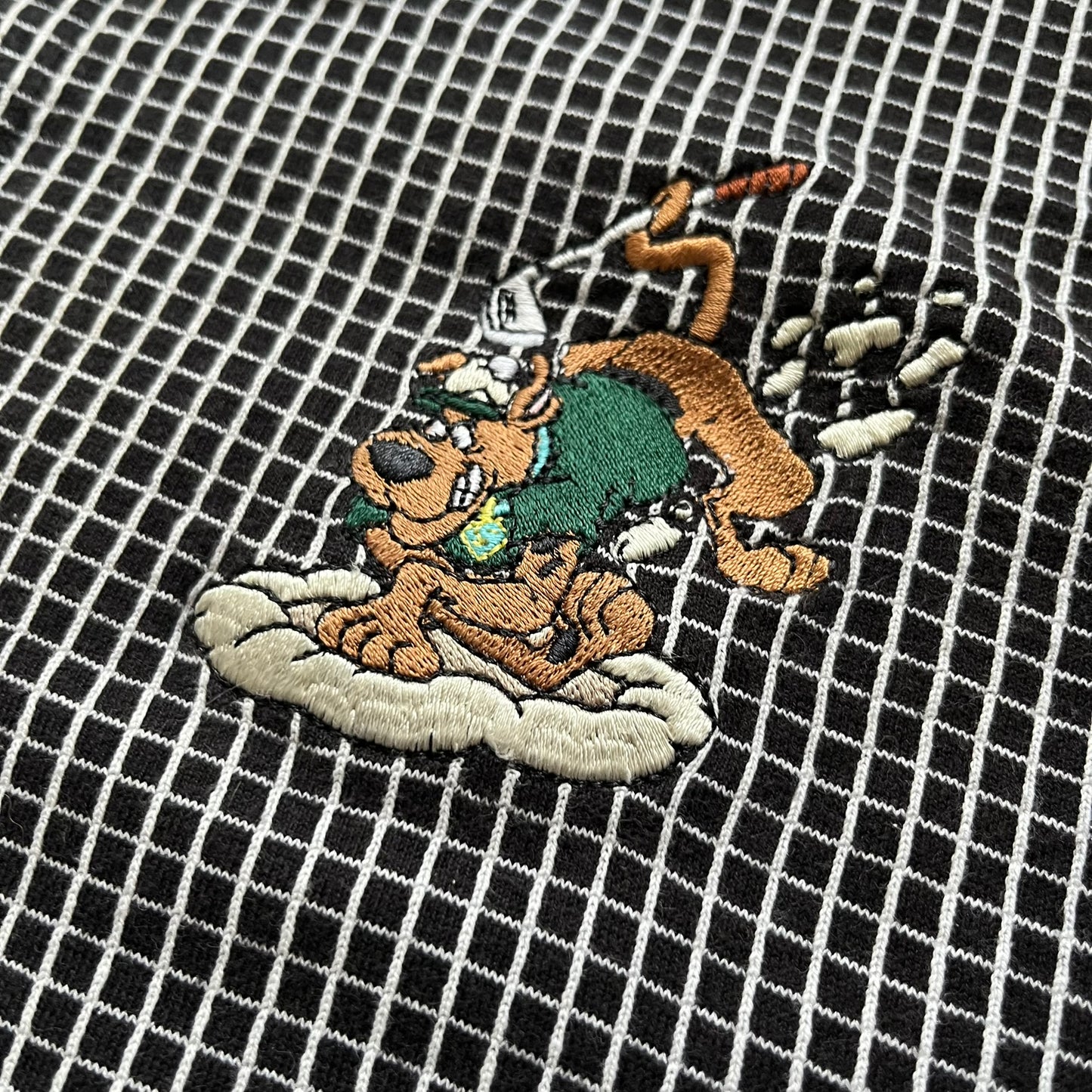 Cartoon Network Scooby-Doo 1999 Golf Polo is a black polo shirt featuring a grid pattern of thin white lines forming small squares across the fabric. The left chest displays an embroidered graphic of Scooby-Doo in a golf outfit digging in sand. The neck tag includes Warner Bros. and Cartoon Network 1999 licensing.
