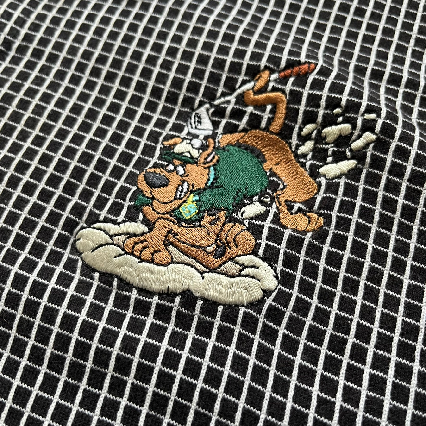 Cartoon Network Scooby-Doo 1999 Golf Polo is a black polo shirt featuring a grid pattern of thin white lines forming small squares across the fabric. The left chest displays an embroidered graphic of Scooby-Doo in a golf outfit digging in sand. The neck tag includes Warner Bros. and Cartoon Network 1999 licensing.