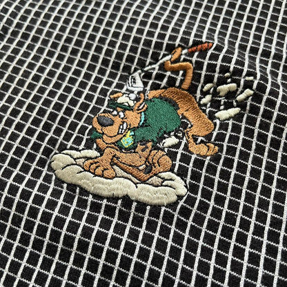 Cartoon Network Scooby-Doo 1999 Golf Polo is a black polo shirt featuring a grid pattern of thin white lines forming small squares across the fabric. The left chest displays an embroidered graphic of Scooby-Doo in a golf outfit digging in sand. The neck tag includes Warner Bros. and Cartoon Network 1999 licensing.