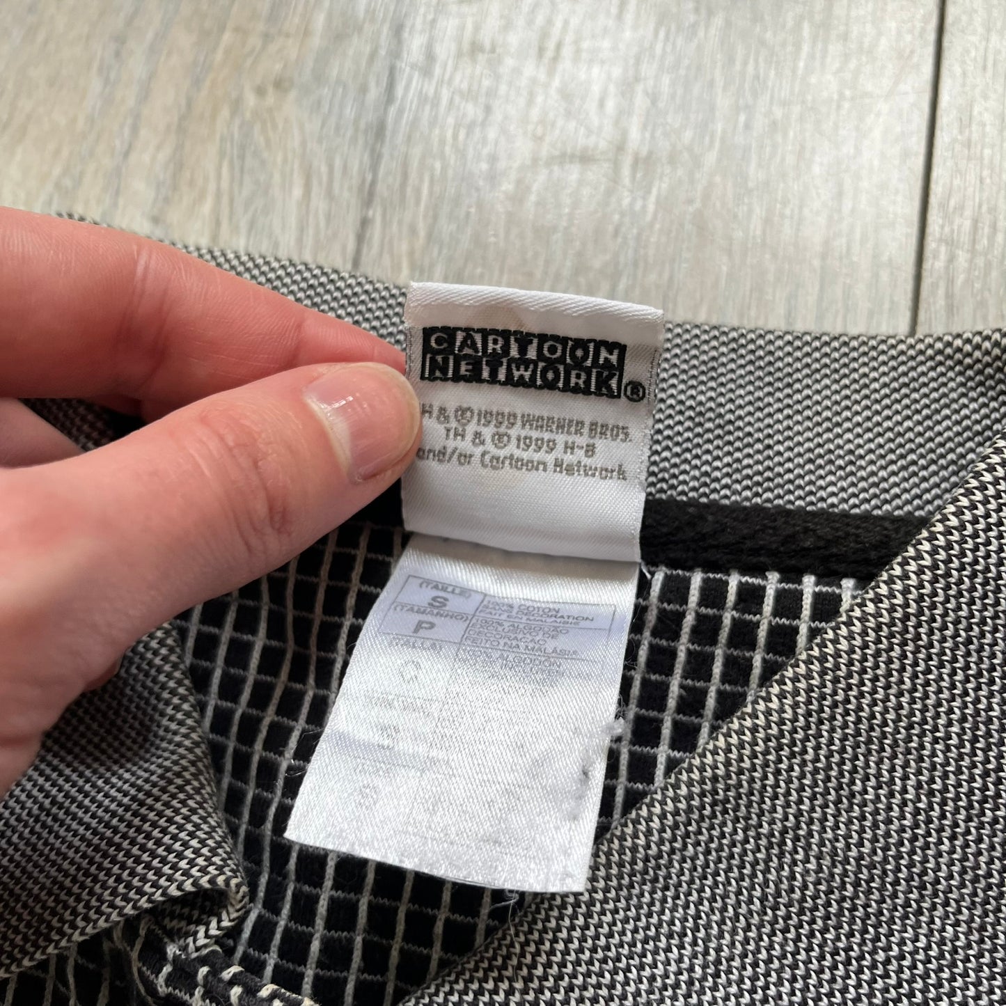 Cartoon Network Scooby-Doo 1999 Golf Polo is a black polo shirt featuring a grid pattern of thin white lines forming small squares across the fabric. The left chest displays an embroidered graphic of Scooby-Doo in a golf outfit digging in sand. The neck tag includes Warner Bros. and Cartoon Network 1999 licensing.