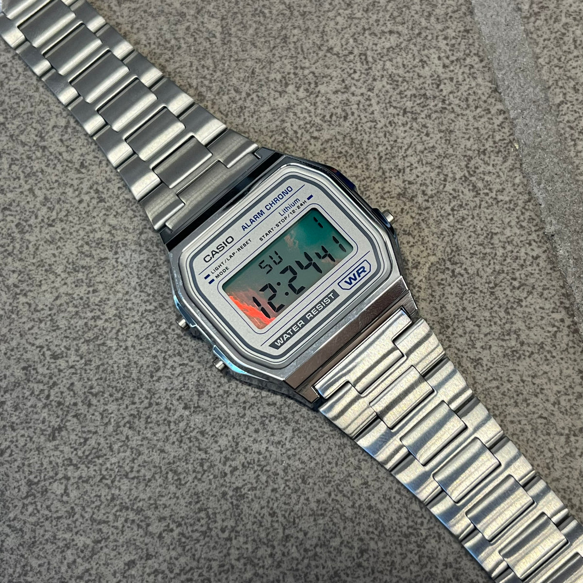 Custom Casio A158 "Chrome" Silver Digital Watch (A158WEA-7EF) is a custom Casio digital watch with a reflective silver filter over the square resin glass screen and a stainless steel band with an adjustable clasp, compatible with wrist sizes from 150 to 205 mm. It has a 1/100-second stopwatch, a daily alarm, an hourly time signal, an auto-calendar, a 12/24-hour format, a green LED light, and water resistance.