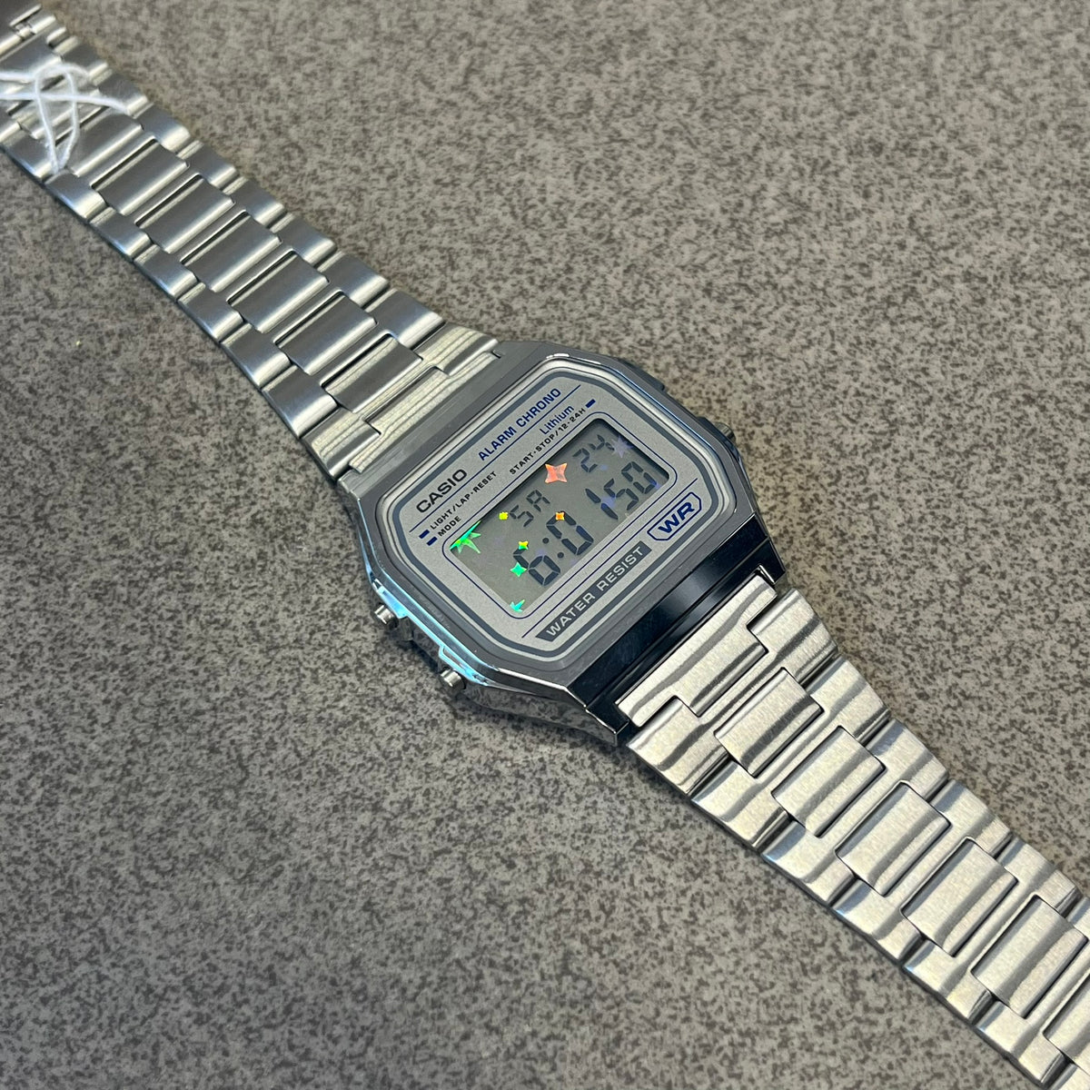 Custom Casio A158 "Double Holo" Silver Digital Watch (A158WEA-7EF) is a custom Casio digital watch with a Double Holo (Stars and normal holo) filter over the square resin glass screen and a stainless steel band with an adjustable clasp, compatible with wrist sizes from 150 to 205 mm. It has a 1/100-second stopwatch, a daily alarm, an hourly time signal, an auto-calendar, a 12/24-hour format, a green LED light, and water resistance.