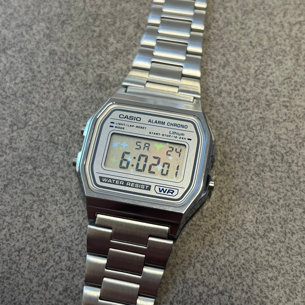 Custom Casio A158 "Double Holo" Silver Digital Watch (A158WEA-7EF) is a custom Casio digital watch with a Double Holo (Stars and normal holo) filter over the square resin glass screen and a stainless steel band with an adjustable clasp, compatible with wrist sizes from 150 to 205 mm. It has a 1/100-second stopwatch, a daily alarm, an hourly time signal, an auto-calendar, a 12/24-hour format, a green LED light, and water resistance.
